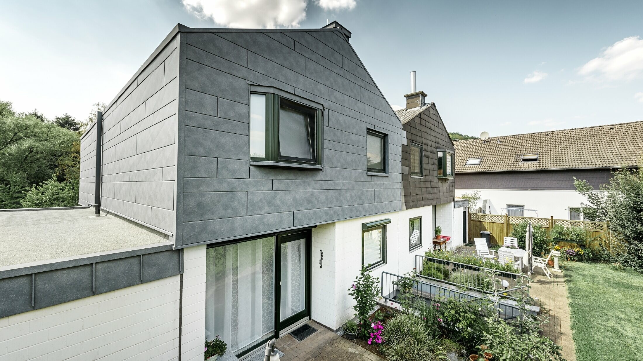 Het bovenste gebouwgedeelte van de helft van de twee-onder-een-kap werd gerenoveerd met het PREFA gevelelement Siding.X in P.10 steengrijs. Meteen daarnaast ter vergelijking de helft van het huis voorafgaand aan de renovatie.