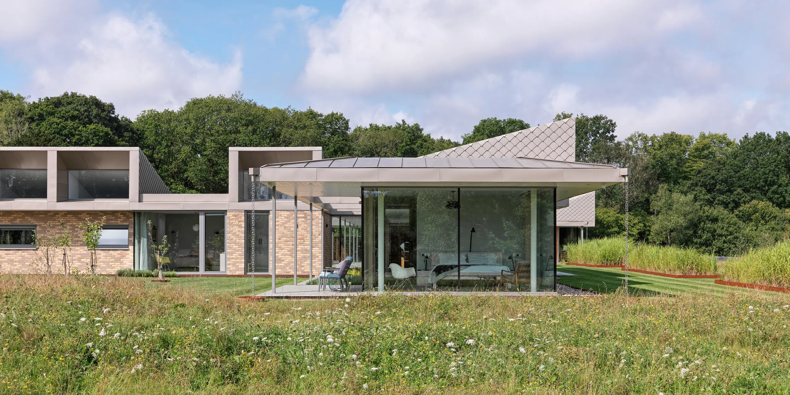 Seitenansicht des modernen Bungalows in West Sussex mit PREFALZ Aluminiumdach und akzentuierten Fassadenbereichen aus PREFA Wandrauten 20 × 20 in P.10 Bronze. Die raumhohen Glasfronten der Wohnbereiche öffnen sich zur Wildblumenwiese im Vordergrund und schaffen eine nahtlose Verbindung zwischen Innenraum und Natur. Das langlebige PREFA Dach- und Fassadensystem verleiht dem Gebäude nicht nur Schutz, sondern auch architektonische Eleganz.
