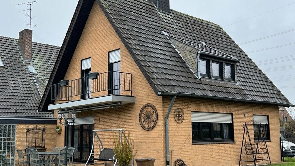 Eengezinswoning van gele baksteen met steil zadeldak en donkere dakbedekking vóór de dakrenovatie; aan de tuinzijde met balkon, dakkapel, terras met zitmeubilair en gazon op de voorgrond.