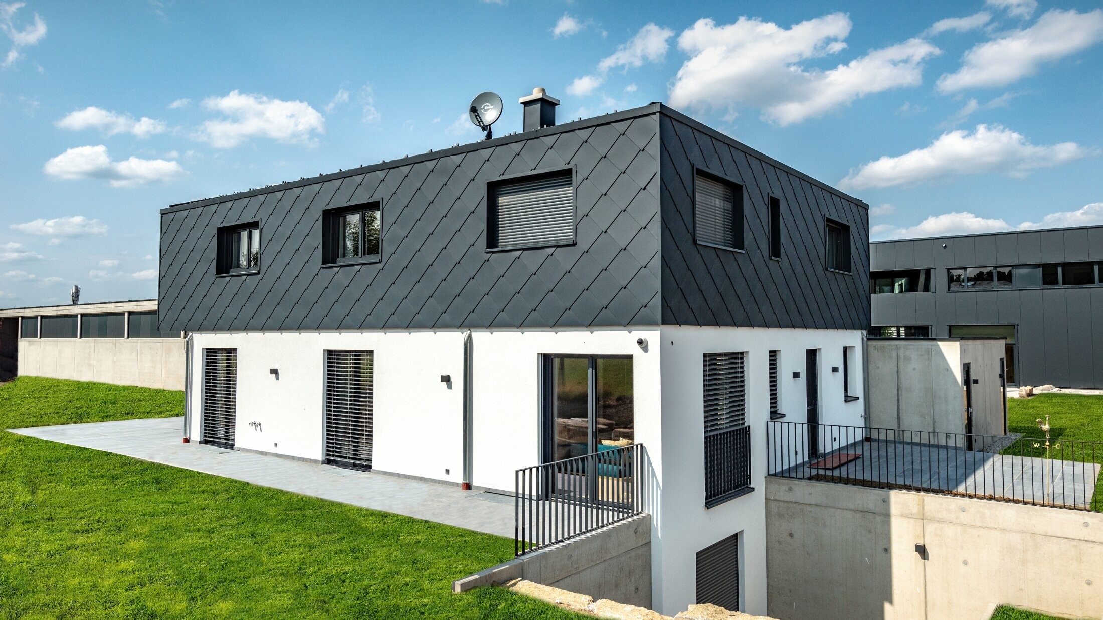 Eenvoudige nieuwbouw met plat dak, de begane grond is wit gestuct, de bovenverdieping is bekleed met de grote PREFA gevellosange in antraciet.