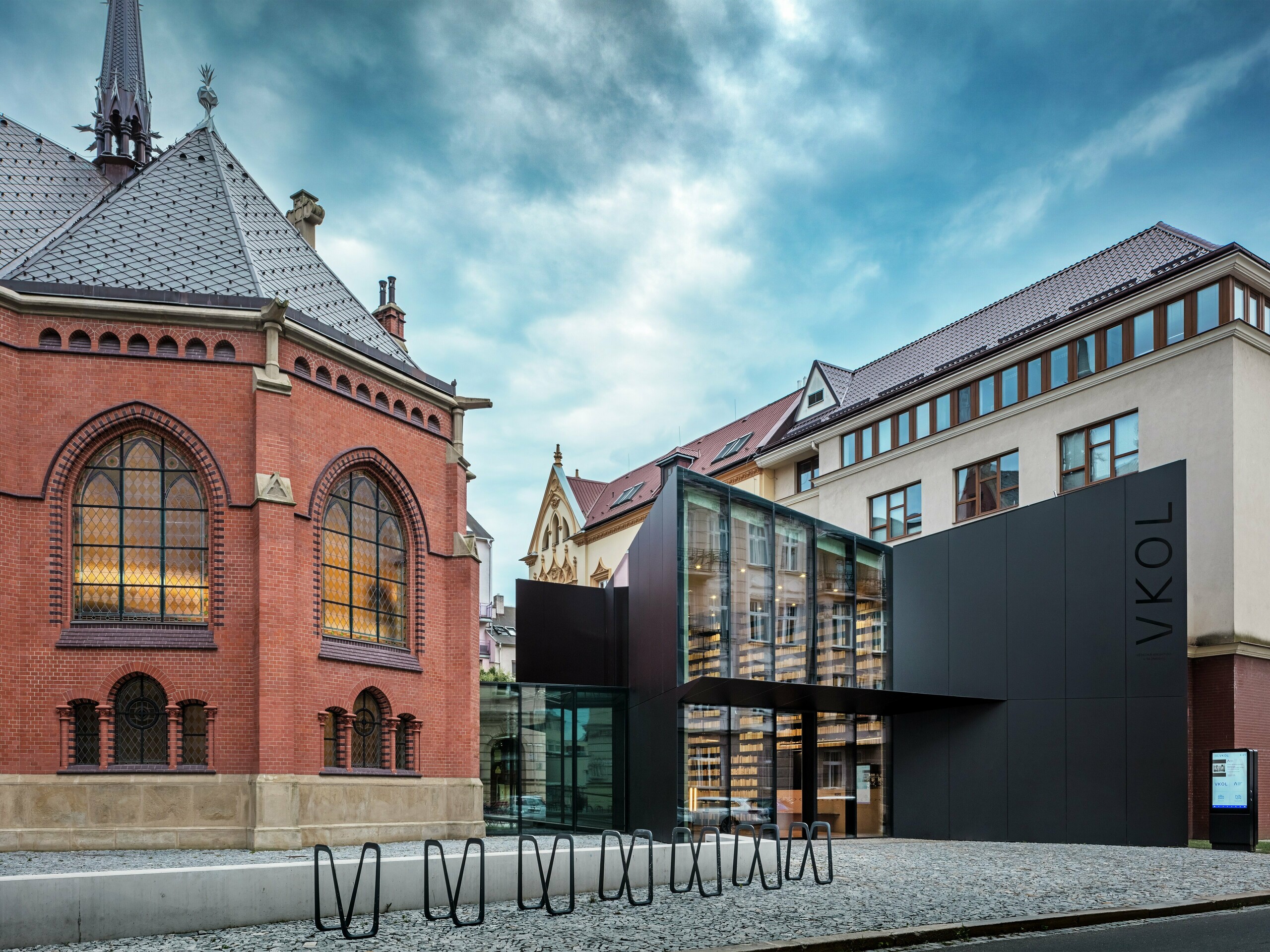 Das Bild zeigt eine faszinierende Verbindung von historischer und zeitgenössischer Architektur in Olmütz. Auf der linken Seite steht die majestätische Rote Kirche mit ihren charakteristischen gotischen Fenstern und roten Backsteinmauern, während der Neubau auf der rechten Seite mit seiner modernen schwarzen PREFABOND Aluminium-Verbundplatten-Fassade kontrastiert. Die gläserne Front des Neubaus gewährt einen Einblick in die innovative Bibliotheksgestaltung und verleiht dem Ensemble einen eleganten, offenen Charakter. Die präzise Verarbeitung der PREFABOND-Platten sorgt für ein einheitliches Erscheinungsbild, das sich harmonisch in die historische Umgebung einfügt und gleichzeitig ein modernes Statement setzt. Die Architektur dieses Projekts verbindet perfekt Tradition und Innovation, wobei die Materialien nicht nur ästhetisch überzeugen, sondern auch für Langlebigkeit und Nachhaltigkeit sorgen.