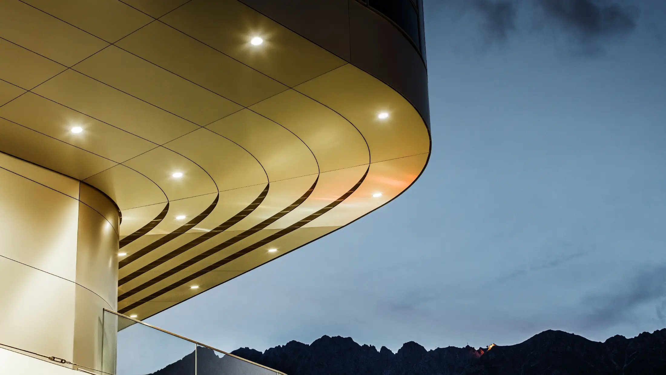 De afbeelding toont een indrukwekkend architectonisch detail van een PREFA PEMA composietplaat van geborsteld aluminium. De plaat is licht gebogen en loopt in elegante, vloeiende lijnen, die 's avonds door gerichte verlichting extra goed tot hun recht komen. De heldere structuur en de edele aluminiumlook van de plaat vormen een aanvulling op het moderne ontwerp van de gevel en bieden een elegante, eigentijdse oplossing voor hoogwaardige architectuur.