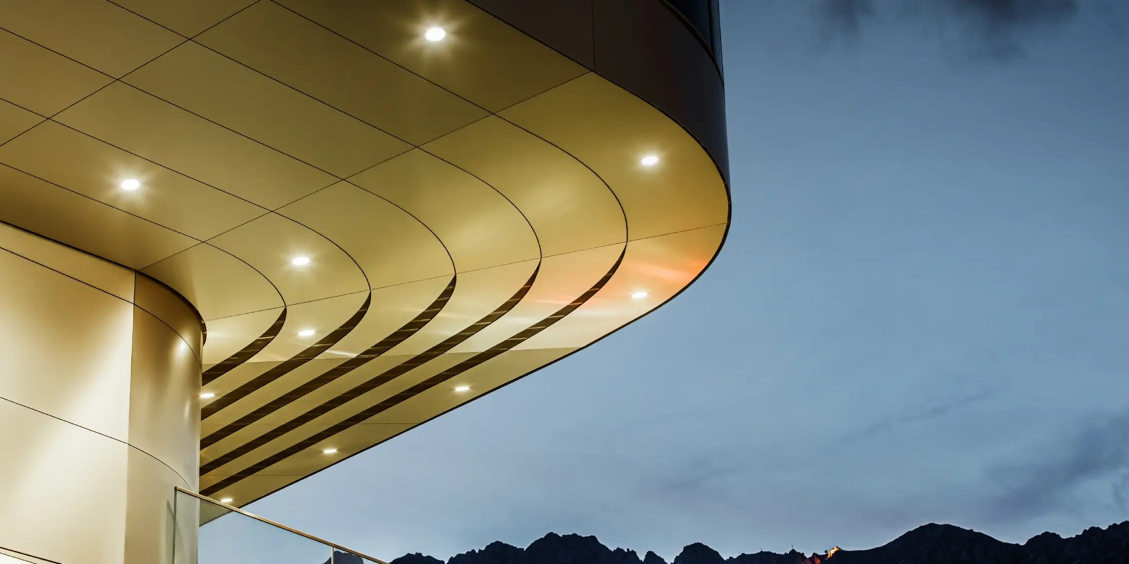 De afbeelding toont een indrukwekkend architectonisch detail van een PREFA PEMA composietplaat van geborsteld aluminium. De plaat is licht gebogen en loopt in elegante, vloeiende lijnen, die 's avonds door gerichte verlichting extra goed tot hun recht komen. De heldere structuur en de edele aluminiumlook van de plaat vormen een aanvulling op het moderne ontwerp van de gevel en bieden een elegante, eigentijdse oplossing voor hoogwaardige architectuur.