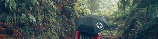 Afbeelding in het bos met wandelaar in rode jas met PREFA paraplu en sporttas, symboliseert zowel PREFA milieubescherming en duurzaamheid als circulaire economie en recycling