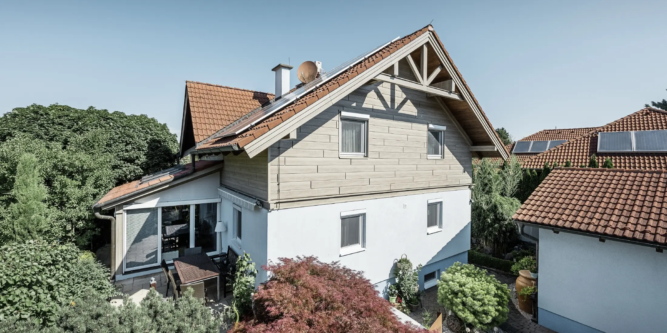 Eengezinswoning met zadeldak en rode dakbedekking, gecombineerd met een moderne gevelontwerp. De bovenste gevel is bekleed met beige-grijze aluminium gevelpaneelen van PREFA, terwijl de begane grond een witte gepleisterde gevel heeft. Op het dak bevinden zich solarmodule. De woning ligt in een groene tuin met terras, tuinmeubilair en weelderige beplanting, omgeven door andere woningen met een vergelijkbare dakarchitectuur.