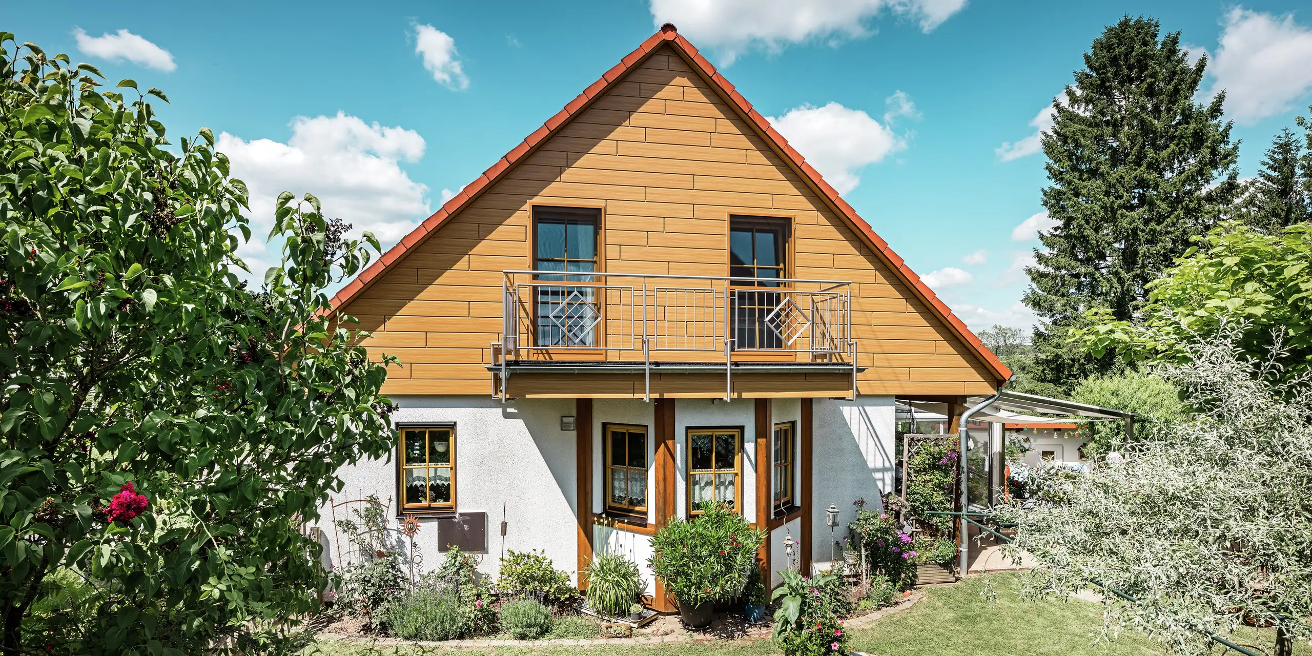 Op deze foto straalt de gerenoveerde gevel van een eengezinswoning in Blieskastel in een warme, natuurlijke eiken naturel-kleur dankzij de PREFA Siding. De hoogwaardige aluminium gevel met realistische houtlook zorgt voor een sterk visueel accent en sluit indrukwekkend aan bij het rode pannendak en de weelderige tuin. Omgeven door bloemen, struiken en weelderig groen oogt het huis uitnodigend en verzorgd. Het PREFA-oppervlak combineert een natuurlijke uitstraling met functionele voordelen zoals hoge weersbestendigheid, stabiliteit en een vrijwel onderhoudsvrije levensduur – een esthetisch hoogtepunt met duurzame meerwaarde.