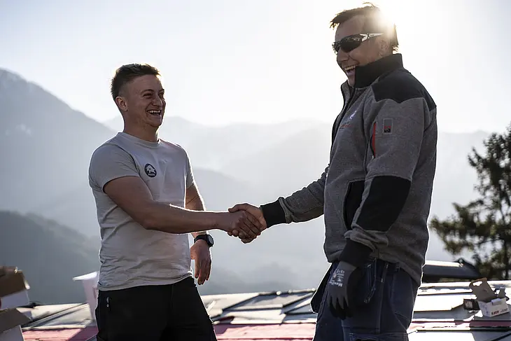 Auf einem geneigten Hausdach stehen ein PREFA-Trainer und ein Spengler, die sich die Hand geben. Beide tragen professionelle Arbeitskleidung und Sicherheitsausrüstung. Im Hintergrund sind eine tiefstehende Sonne und Verpackungsschachteln von PREFA-Aluminiumprodukten zu sehen. Diese Szene symbolisiert eine erfolgreiche Zusammenarbeit und einen fachlichen Austausch mit Handschlagqualität. Der PREFA-Trainer unterstützt den Spengler mit seinem Know-how zu den Themen Verarbeitung, Technik und Produktsysteme. Das Bild steht für die enge Betreuung von Handwerkern durch PREFA – von der Schulung bis zur professionellen Umsetzung auf der Baustelle.