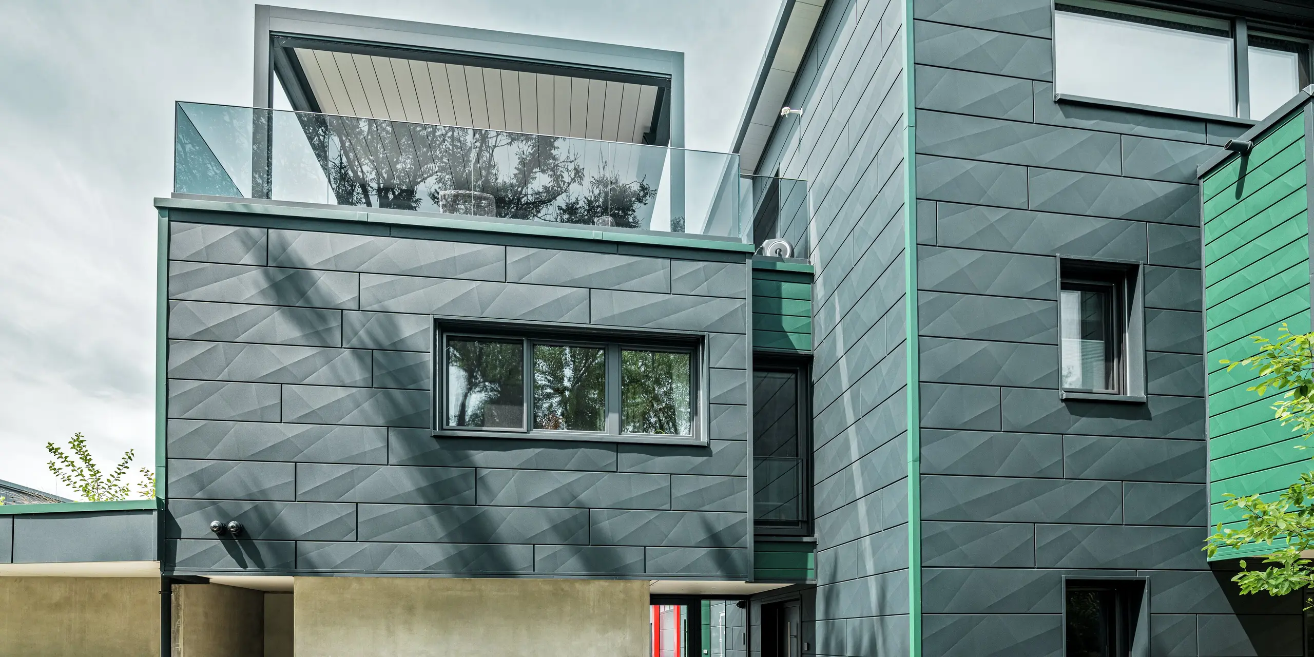 Das moderne Einfamilienhaus in Türkheim beeindruckt mit seiner einzigartigen Fassade aus PREFA Siding X-Paneelen in den Farben P.10 Anthrazit und P.10 Moosgrün. Die Längs- und Querkantungen der Aluminiumpaneele verleihen dem Gebäude eine dynamische Optik. Die Perspektive zeigt das Haus von der Gartenseite aus mit Blick auf den Eingangsbereich. Über dem Eingangsbereich ist eine Dachterrasse mit einer modernen Pergola zu erkennen.