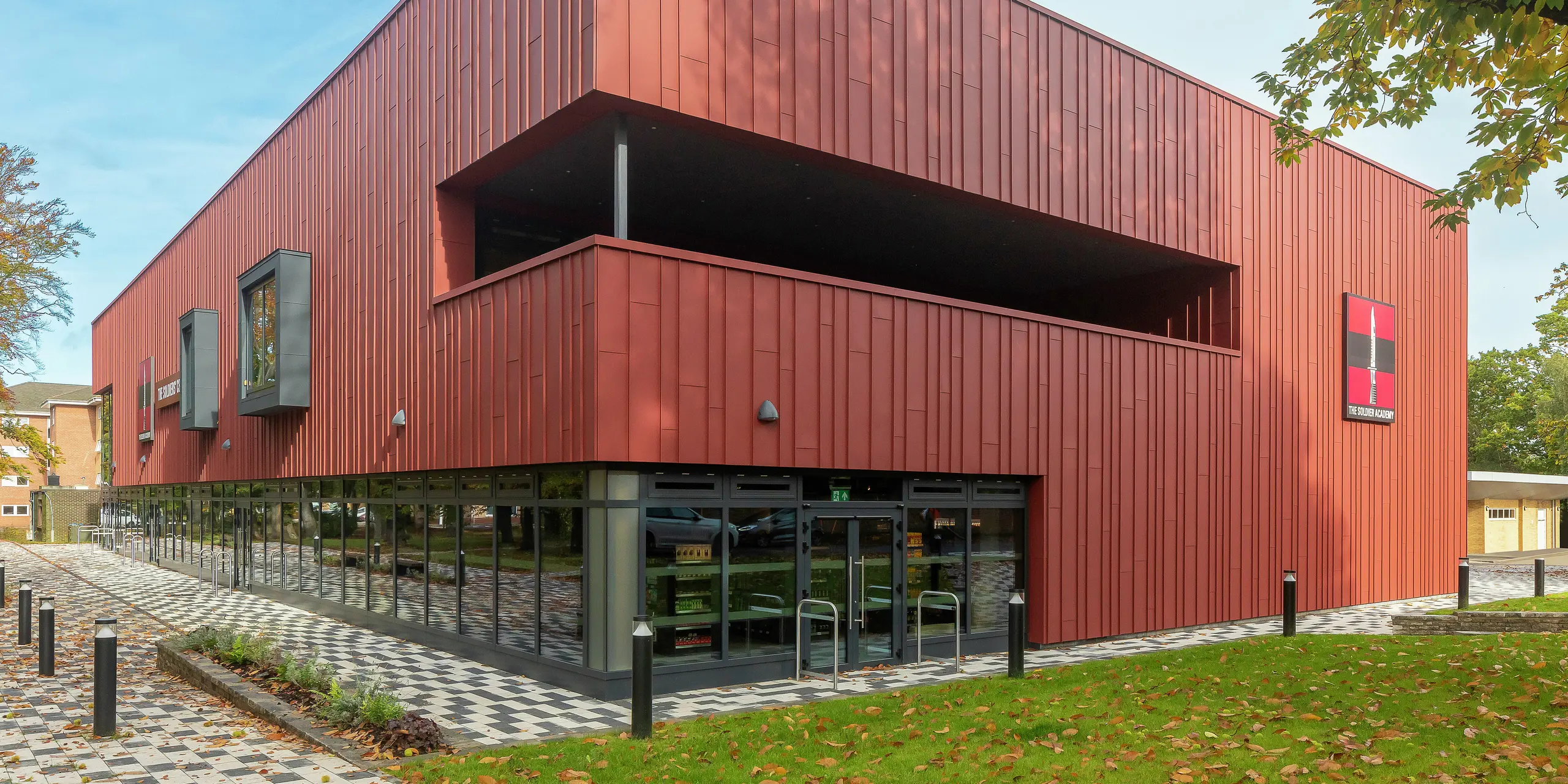 Zeitgemäße Architektur mit PREFALZ: Das Soldiers' Centre in Pirbright überzeugt mit einer modernen Stehfalzfassade aus PREFALZ in P.10 Oxydrot. Die großflächige Verglasung im Erdgeschoss schafft Transparenz und Offenheit, während Fensterrahmen in P.10 Anthrazit stilvolle Akzente setzen. Ein nachhaltiges Baukonzept mit langlebigen PREFA Aluminiumprodukten für höchste Design- und Qualitätsansprüche.