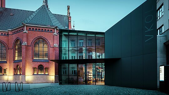 Das Bild zeigt die beeindruckende Verschmelzung von historischer und moderner Architektur in Olmütz. Die markante Rote Kirche mit ihrem gotischen Stil, einschließlich der kunstvollen Fenster und des hohen Turms, wird durch den modernen Neubau mit seiner minimalistischen, schwarzen Fassade aus PREFABOND Aluminium-Verbundplatten ergänzt. Die transparenten Glasflächen des Neubaus reflektieren das historische Umfeld und bieten einen faszinierenden Kontrast zur soliden Struktur der Kirche. Die präzise Verarbeitung der modernen Materialien, gepaart mit der harmonischen Verbindung von Alt und Neu, verleiht dem Gebäude eine zeitgemäße Eleganz, während es gleichzeitig die Geschichte der Umgebung respektiert. Diese Kombination aus innovativen und langlebigen Materialien wie PREFABOND gewährleistet sowohl ästhetische Schönheit als auch Funktionalität.