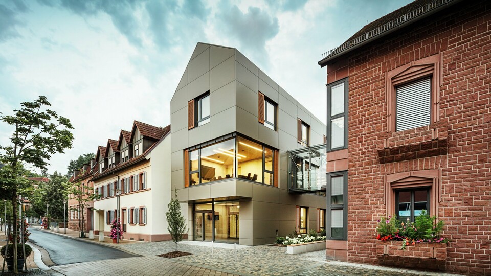 Das Bild zeigt das moderne Rathaus in Alzenau, dessen Fassade mit PREFA-Aluminiumverbundplatten in einem warmen Bronzeton verkleidet ist. Große Glasflächen unterbrechen die Fassade und geben den Blick auf ein lichtdurchflutetes Interieur frei. Ein gläserner Verbindungsgang auf der rechten Bildseite verbindet das Gebäude mit einem nahegelegenen traditionellen Backsteingebäude und schafft so einen interessanten Kontrast zwischen moderner und historischer Architektur.