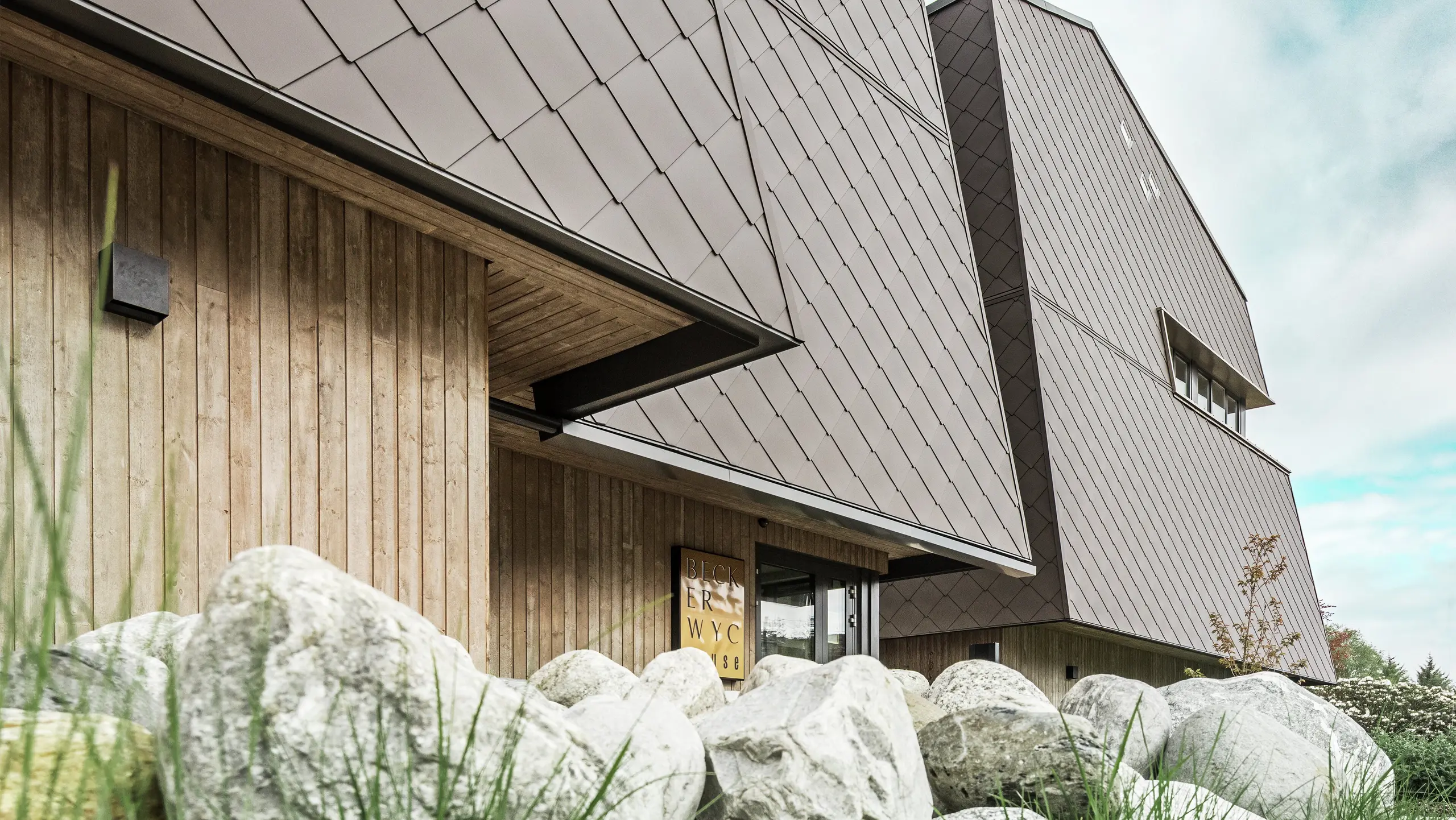 Zijaanzicht van een hotel, de gevel van de begane grond is verticaal bekleed met donker hout. Daarboven is een gevelsprong, vanaf hier is de gevel bekleed met aluminium panelen in walnootbruin.