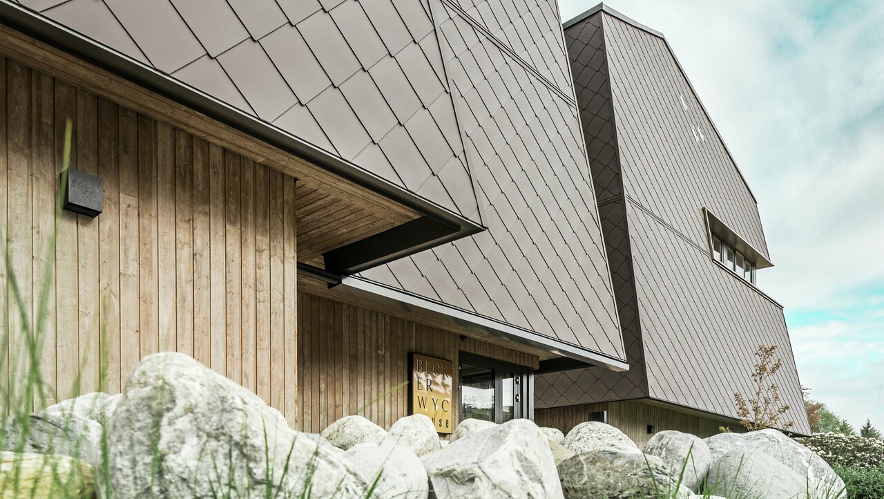 Zijaanzicht van een hotel, de gevel van de begane grond is verticaal bekleed met donker hout. Daarboven is een gevelsprong, vanaf hier is de gevel bekleed met aluminium panelen in walnootbruin.
