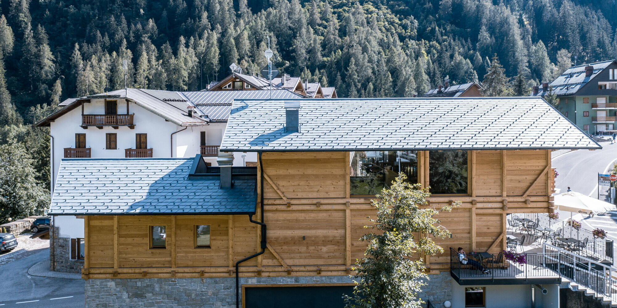 Seitenansicht eines gemütlichen Après-Ski-Restaurants mit einer Außenterrasse, umrahmt von atemberaubenden alpinen Wäldern. Die Fassade kombiniert natürliches Holz mit robustem Stein, ergänzt durch hochwertige PREFA Aluminiumdachschindeln DS.19 in P.10 Steingrau, die perfekt in die malerische Bergkulisse Italiens integriert sind.