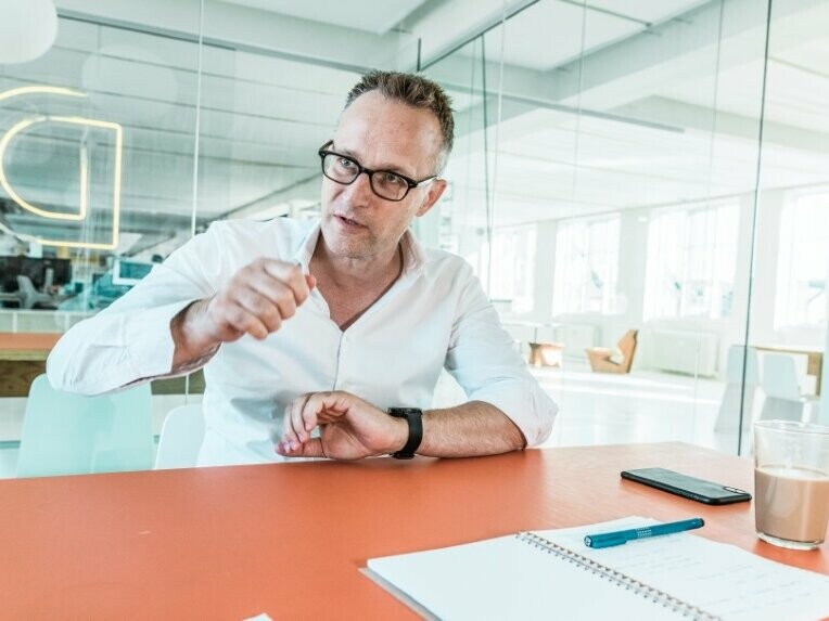 Architekt Noel Wibrand sitzt mit dem Gesicht zur Kamera an einem Schreibtisch und erklärt etwas