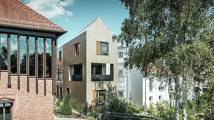 Translate to Dutch: Das Bild zeigt das moderne Rathaus in Alzenau, dessen Fassade aus Aluminium-Verbundplatten von PREFA in Bronze besteht. Der spitze Dachbereich und die klar strukturierten Fenster mit Holzjalousien verleihen dem Gebäude eine elegante und zeitgenössische Ästhetik. Im Vordergrund ist ein grüner Bereich mit einem gepflasterten Weg und Bäumen zu sehen, der das urbane Design des Gebäudes harmonisch ergänzt.