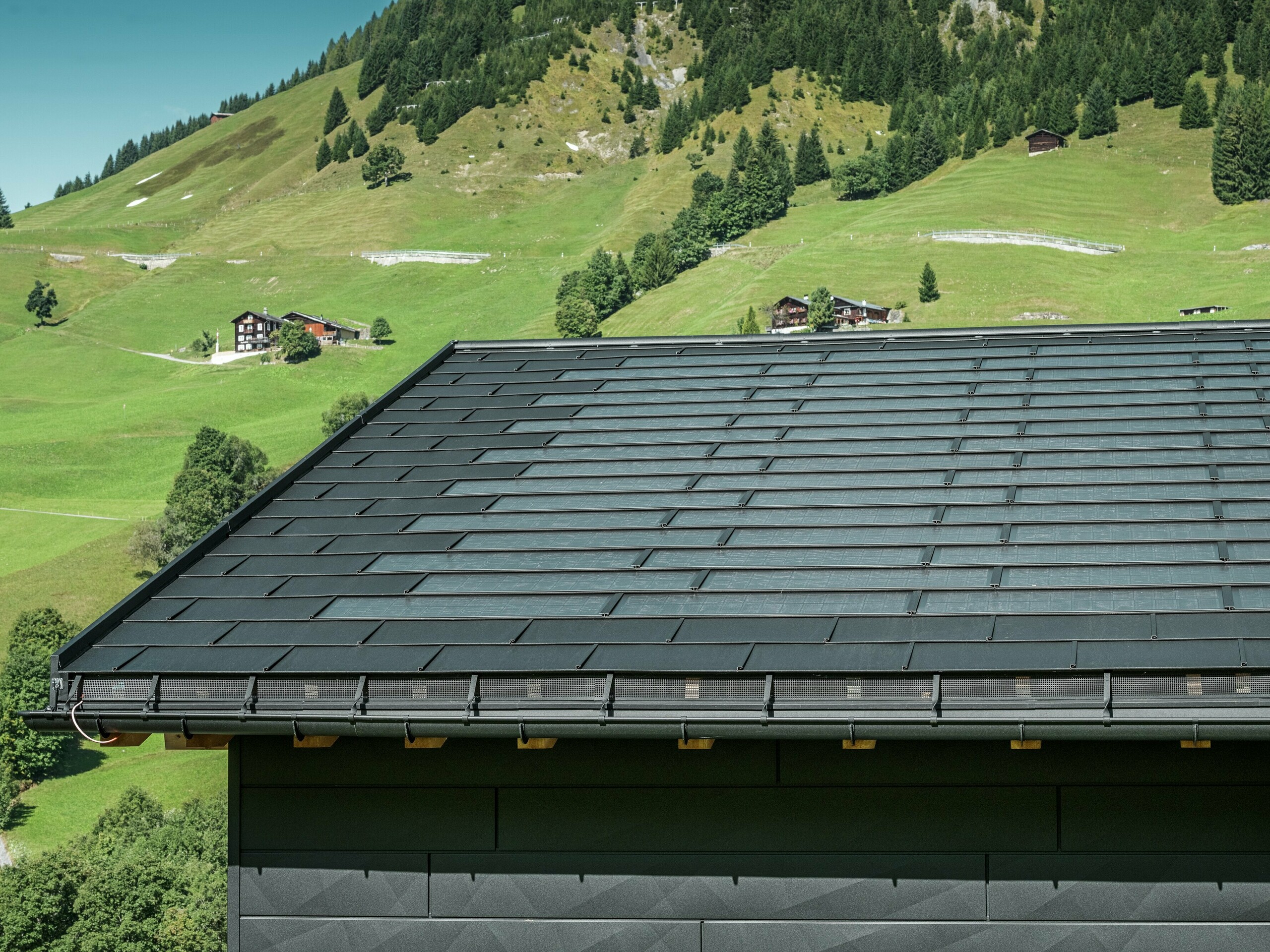 Close-up van de dakrand van een moderne eengezinswoning in de alpenomgeving van Ascharina in Zwitserland. Het dak is bedekt met de innovatieve PREFA solar-dakpan en de traditionele dakpan R.16, beide in P.10 zwart. De PREFA mastgoot en de PREFA afvoerbuis zorgen voor een betrouwbare dakafvoersystemen. Aan de onderkant van de foto zijn de onmiskenbare PREFA Siding.X gevelpanelen te zien. De strakke lijnen van de aluminium producten onderstrepen het moderne design te midden van een schilderachtig berglandschap.