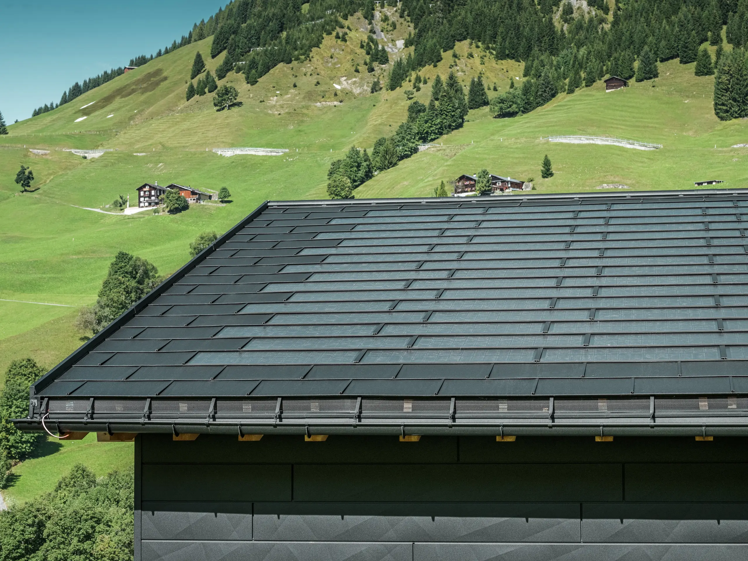 Close-up van de dakrand van een moderne eengezinswoning in de alpenomgeving van Ascharina in Zwitserland. Het dak is bedekt met de innovatieve PREFA solar-dakpan en de traditionele dakpan R.16, beide in P.10 zwart. De PREFA mastgoot en de PREFA afvoerbuis zorgen voor een betrouwbare dakafvoersystemen. Aan de onderkant van de foto zijn de onmiskenbare PREFA Siding.X gevelpanelen te zien. De strakke lijnen van de aluminium producten onderstrepen het moderne design te midden van een schilderachtig berglandschap.