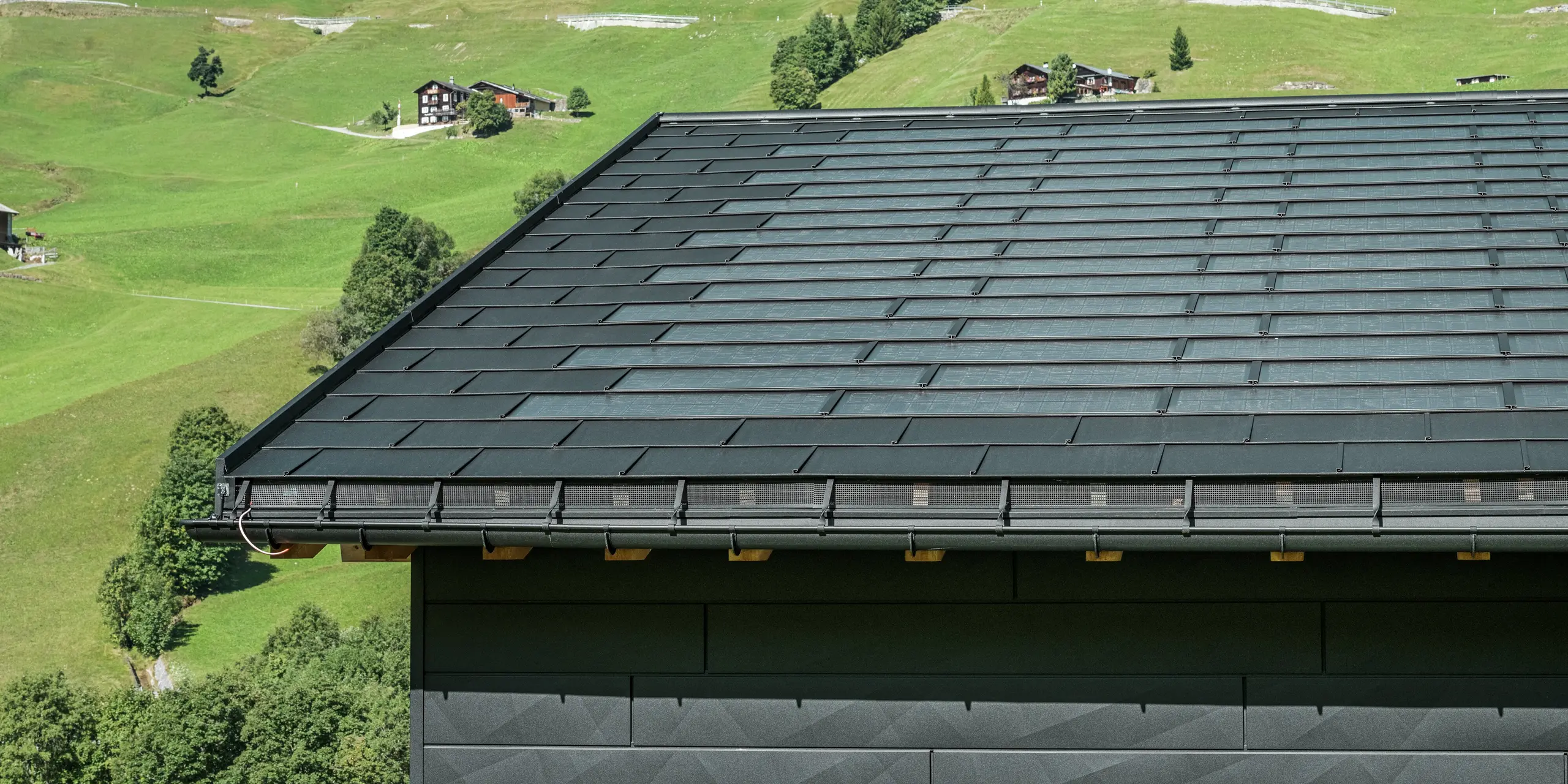 Close-up van de dakrand van een moderne eengezinswoning in de alpenomgeving van Ascharina in Zwitserland. Het dak is bedekt met de innovatieve PREFA solar-dakpan en de traditionele dakpan R.16, beide in P.10 zwart. De PREFA mastgoot en de PREFA afvoerbuis zorgen voor een betrouwbare dakafvoersystemen. Aan de onderkant van de foto zijn de onmiskenbare PREFA Siding.X gevelpanelen te zien. De strakke lijnen van de aluminium producten onderstrepen het moderne design te midden van een schilderachtig berglandschap.