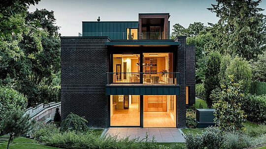 Die Aufnahme zeigt die moderne Rückseite der Budapester Mehrgenerationen‑Villa in der Abenddämmerung. Großzügige Glasfronten öffnen den Blick auf warm erleuchtete Innenräume, während die Kombination aus dunkelroter Backsteinfassade und PREFALZ Aluminium in P.10 Anthrazit klare architektonische Linien betont. Die präzise ausgeführten Winkelstehfalze unterstreichen die hochwertige Verarbeitung und Langlebigkeit der PREFA Aluminiumprodukte. Umgeben von üppigem Grün fügt sich das Gebäude harmonisch in die Gartenlandschaft ein und vermittelt einen eleganten, modernen Charakter.