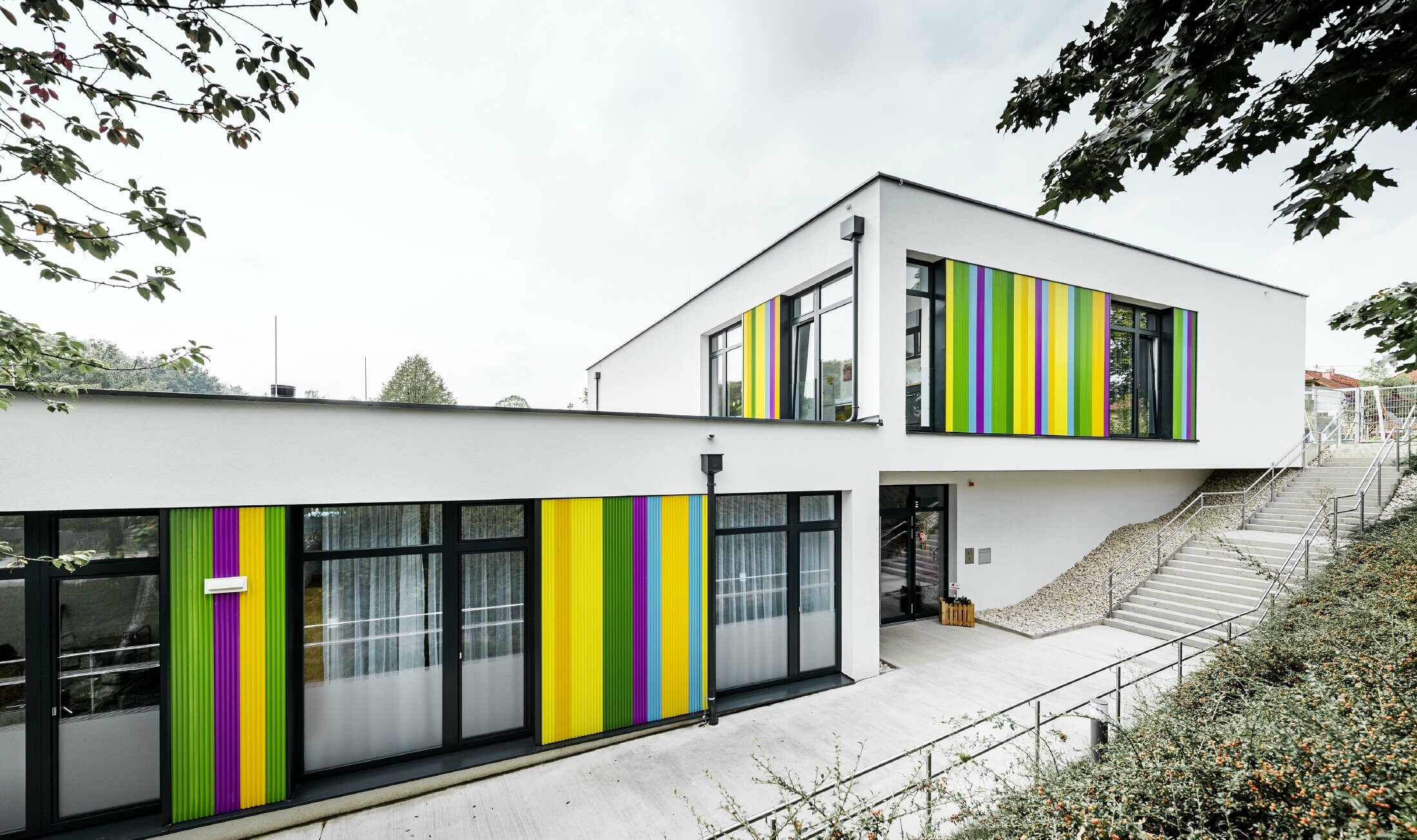 Kleurrijke gevelelementen van PREFA voor de kleuterschool in Hargelsberg. Het gebouw heeft een plat dak en grote ramen tot op de grond.