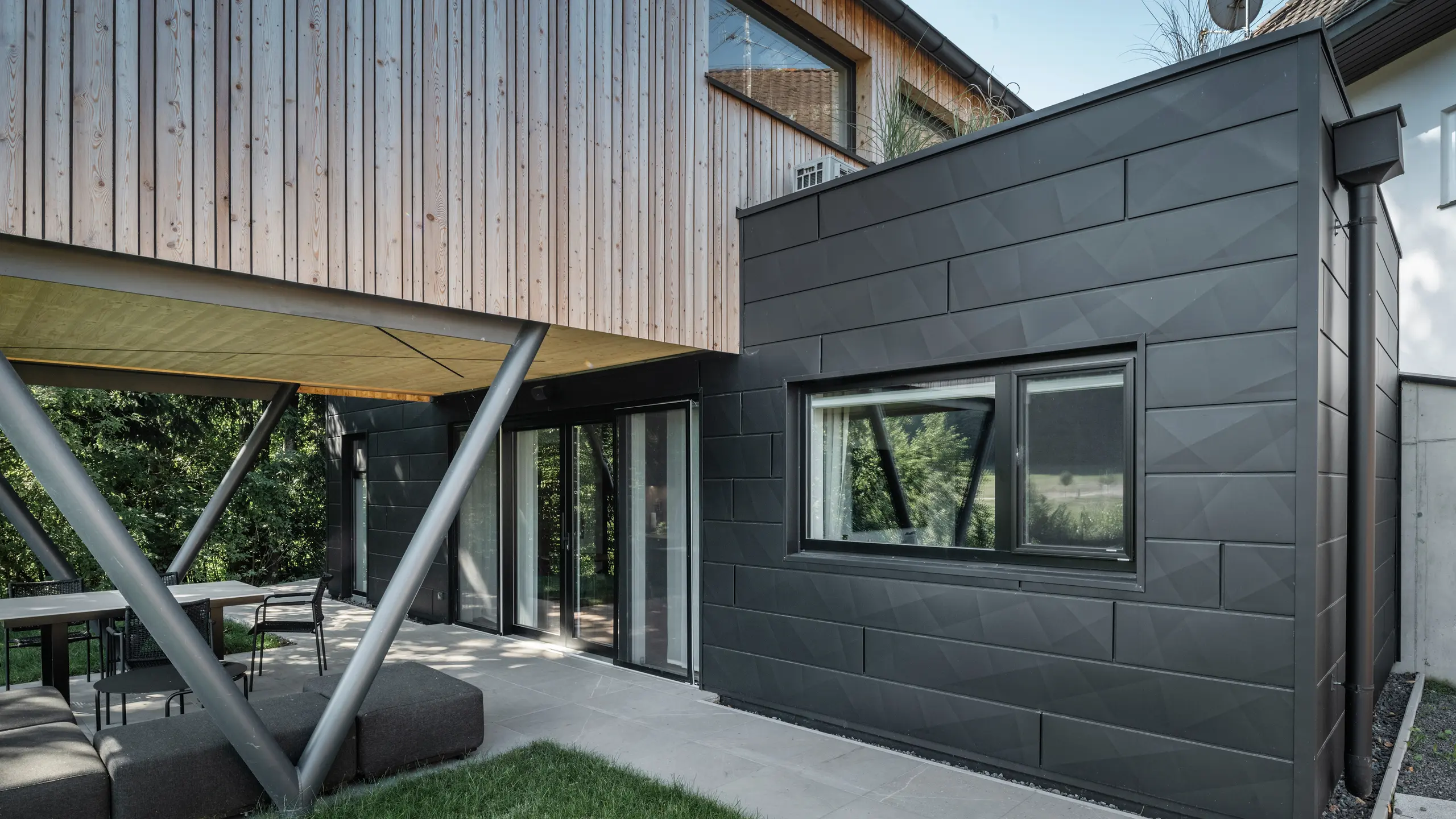Architectonisch hoogtepunt: ‘Woodstock Chalet 5’ in St. Martin am Innkreis, Opper-Oostenrijk. De moderne accommodatie voor bruiloften wordt benadrukt door het gebruik van antracietkleurige PREFA Siding.X-gevelpaneelen. De aluminium elementen vormen een dikte contrast met de lichte houten panelen en de metalen ondersteuningsconstructies. De donkere raamkozijnen passen harmonieus in het gevelontwerp, waardoor het gebouw een elegante uitstraling krijgt. Deze foto toont het esthetische evenwicht tussen de natuurlijke houtstructuur en de industriële charme van de aluminium gevel, die staat voor het ontwerp van eigentijdse gebouwen.
