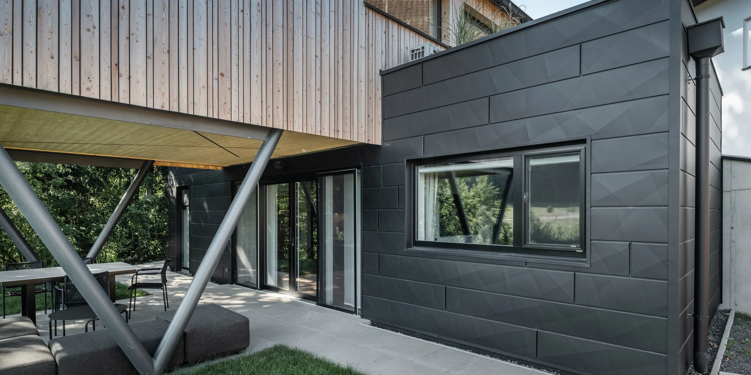 Architectonisch hoogtepunt: ‘Woodstock Chalet 5’ in St. Martin am Innkreis, Opper-Oostenrijk. De moderne accommodatie voor bruiloften wordt benadrukt door het gebruik van antracietkleurige PREFA Siding.X-gevelpaneelen. De aluminium elementen vormen een dikte contrast met de lichte houten panelen en de metalen ondersteuningsconstructies. De donkere raamkozijnen passen harmonieus in het gevelontwerp, waardoor het gebouw een elegante uitstraling krijgt. Deze foto toont het esthetische evenwicht tussen de natuurlijke houtstructuur en de industriële charme van de aluminium gevel, die staat voor het ontwerp van eigentijdse gebouwen.