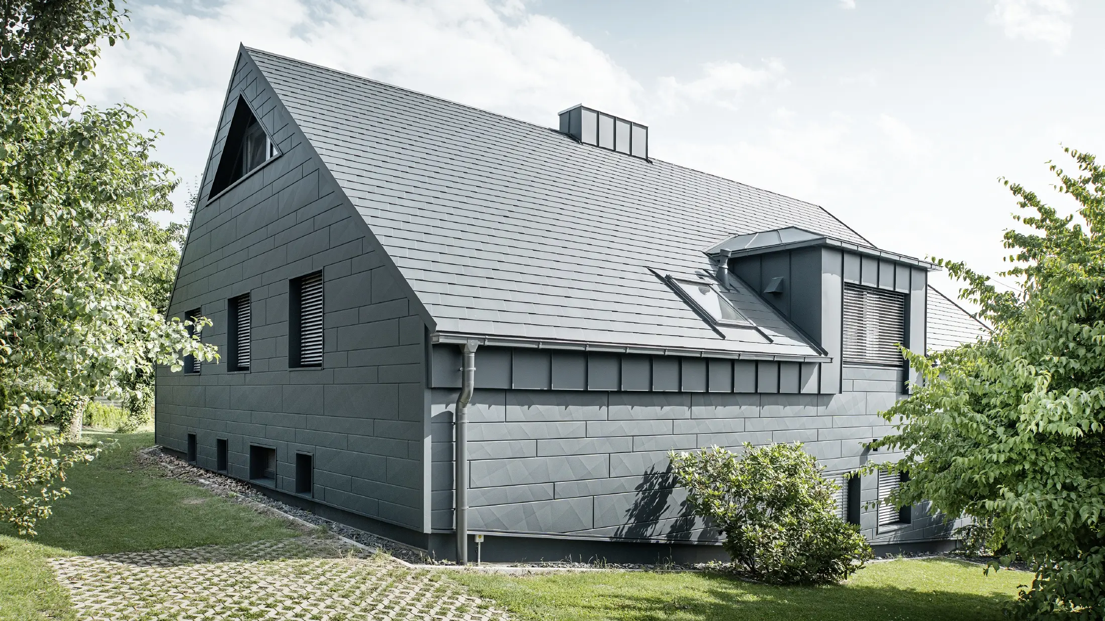 Grote eengezinswoning met zadeldak en veel ramen. Het hele huis is bekleed met een aluminium omhulsel in antraciet, op het dak zijn PREFA aluminium dakspanen gebruikt en op de gevel Siding.X in antraciet.