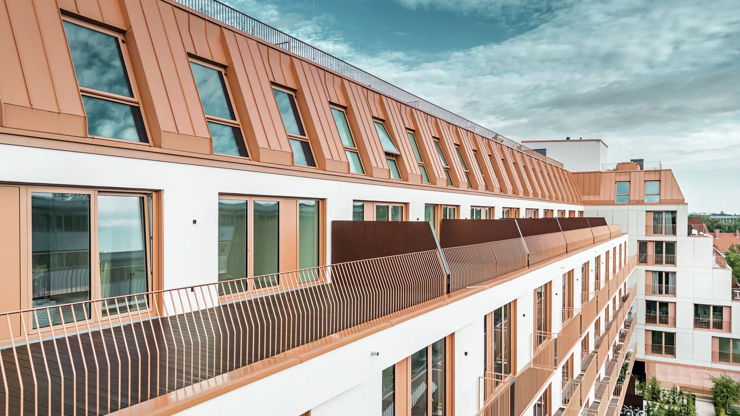 Das moderne Wohngebäude Van B in München besticht durch seine markante Dachlandschaft aus PREFALZ-Aluminium in der Sonderfarbe IGP Orange HDP. Die langlebige und wartungsarme Dachkonstruktion von PREFA gewährleistet ästhetische Integrität und hohe Witterungsbeständigkeit. Die elegante Fassadengestaltung mit kupferroten Fensterrahmen und großzügigen Balkonen unterstreicht das innovative Architekturkonzept. Die ideale Lösung für urbane Bauprojekte mit hohen Ansprüchen an Design und Funktionalität.