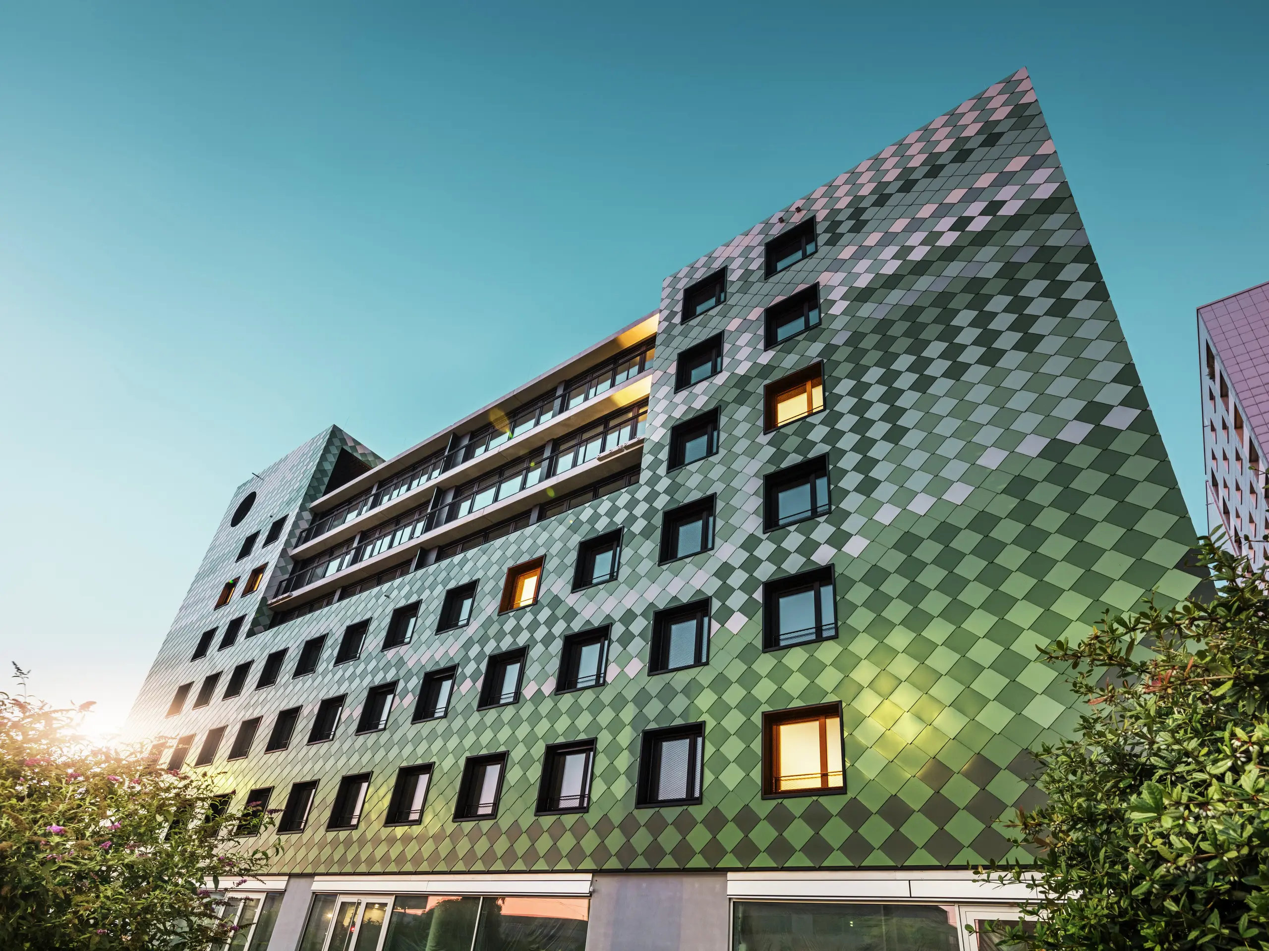 Das moderne Wohn- und Geschäftsgebäude „Pick-Nick“ in Dresden besticht durch seine markante PREFA-Fassade aus Aluminium-Rhombusen in den Sonderfarben Olivgrün, Graugrün, Resedagrün, Opalgrün, Hellelfenbein, Perlweiß und Hellbronze. Die kreative Fassadengestaltung erinnert an ein Reisfeld und erzeugt ein lebendiges Farbenspiel. Die klare Architektur, die schwarzen Fensterrahmen und die Langlebigkeit der PREFA-Materialien prägen das Gebäude. Das tiefstehende Sonnenlicht spiegelt sich auf der Fassade und den Fensterflächen.