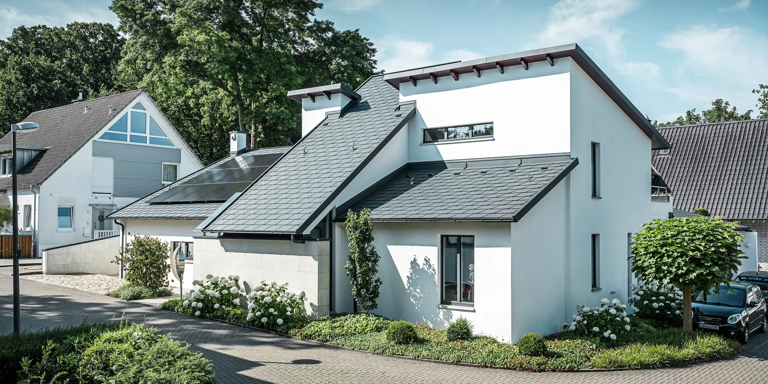 Moderne eengezinswoning met witte gepleisterde gevel en donker PREFA-hellend dak van aluminium. Op het dak zijn geïntegreerde fotovoltaïsche modules en aluminium dakafvoersystemen in antraciet te zien. Het gebouw ligt in een verzorgde woonwijk met beplante voortuinen, een geplaveide oprit en omliggende eengezinswoningen.