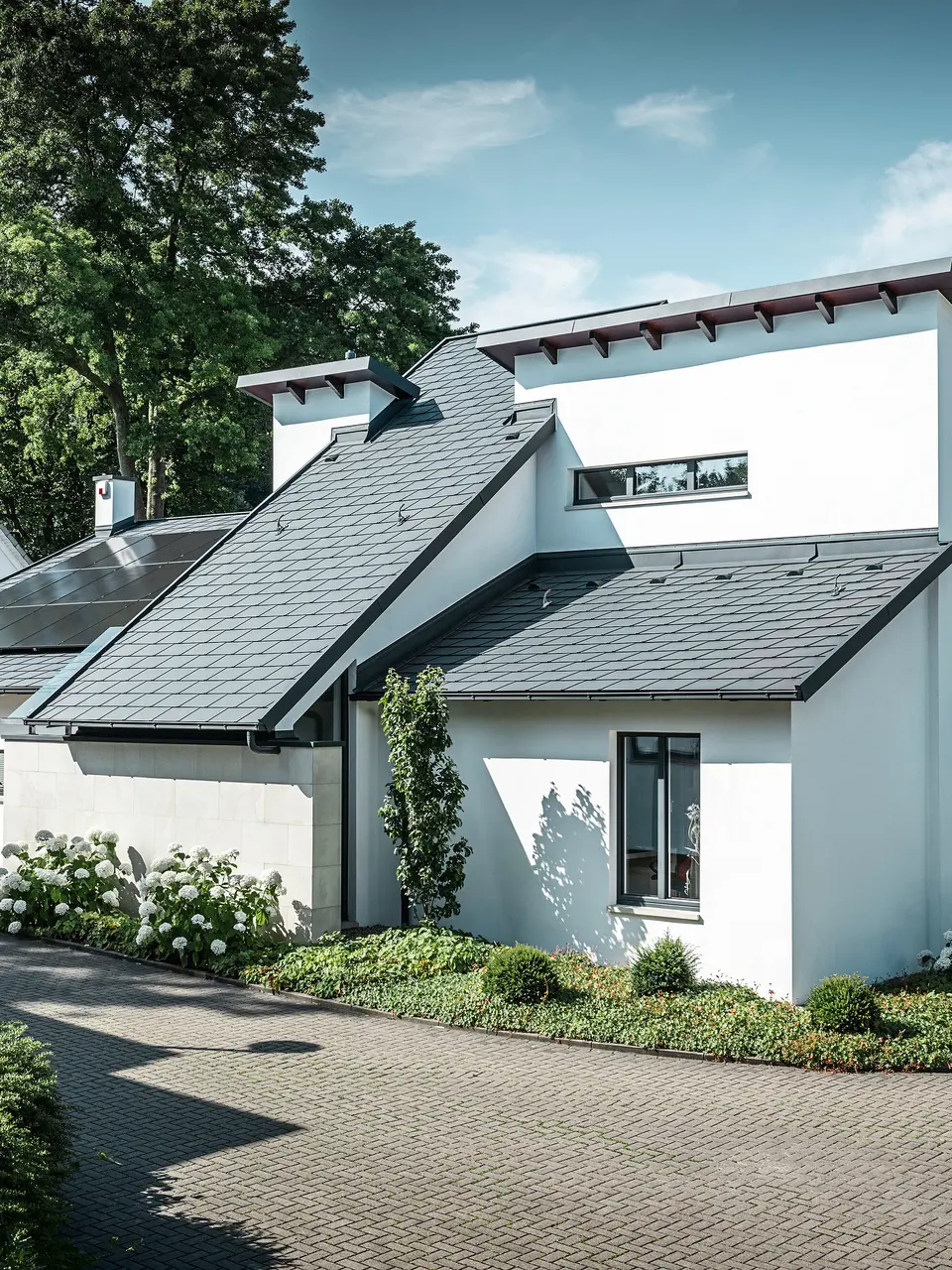 Moderne eengezinswoning met witte gepleisterde gevel en donker PREFA-hellend dak van aluminium. Op het dak zijn geïntegreerde fotovoltaïsche modules en aluminium dakafvoersystemen in antraciet te zien. Het gebouw ligt in een verzorgde woonwijk met beplante voortuinen, een geplaveide oprit en omliggende eengezinswoningen.