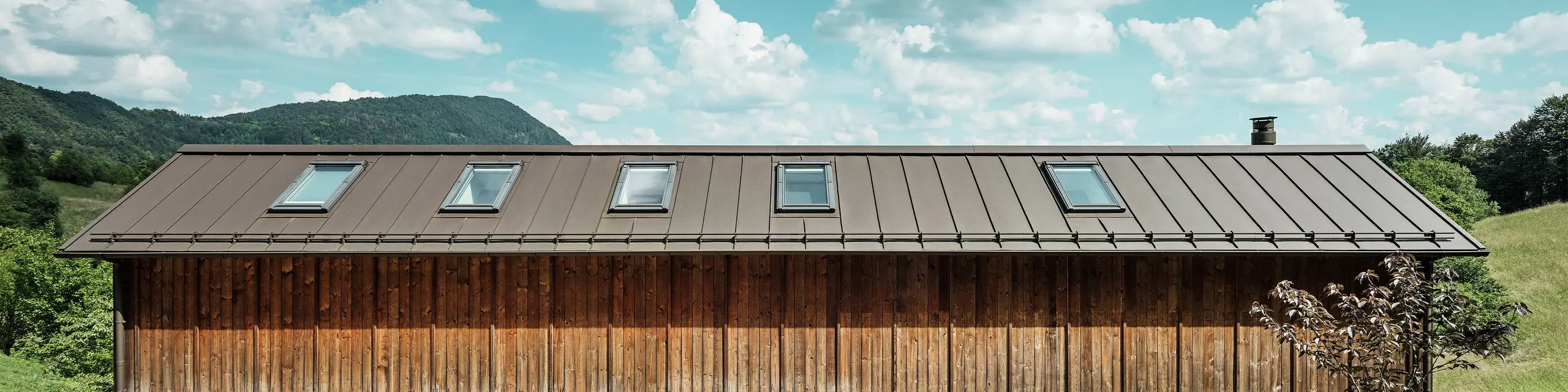Dieses moderne Haus am Hang besticht durch seine minimalistische Holzfassade und das robuste PREFALZ-Aluminiumdach in der Farbe P.10 Braun. Das Stehfalzdach zeichnet sich durch hohe Witterungsbeständigkeit und Langlebigkeit sowie eine harmonische Einbindung in die natürliche Umgebung aus. Integrierte Dachfenster und klare Linien unterstreichen die zeitlose Architektur. Das Bild zeigt den Eingangsbereich dieses kleinen Einfamilienhauses.