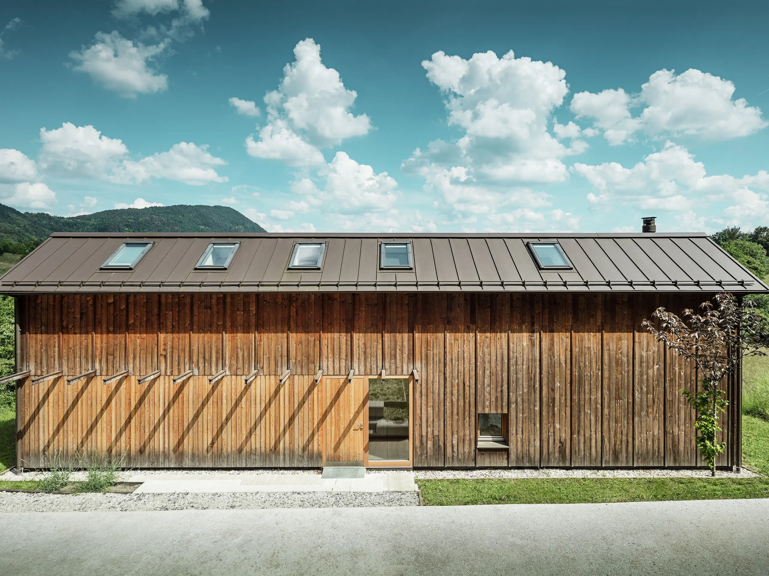 Dieses moderne Haus am Hang besticht durch seine minimalistische Holzfassade und das robuste PREFALZ-Aluminiumdach in der Farbe P.10 Braun. Das Stehfalzdach zeichnet sich durch hohe Witterungsbeständigkeit und Langlebigkeit sowie eine harmonische Einbindung in die natürliche Umgebung aus. Integrierte Dachfenster und klare Linien unterstreichen die zeitlose Architektur. Das Bild zeigt den Eingangsbereich dieses kleinen Einfamilienhauses.