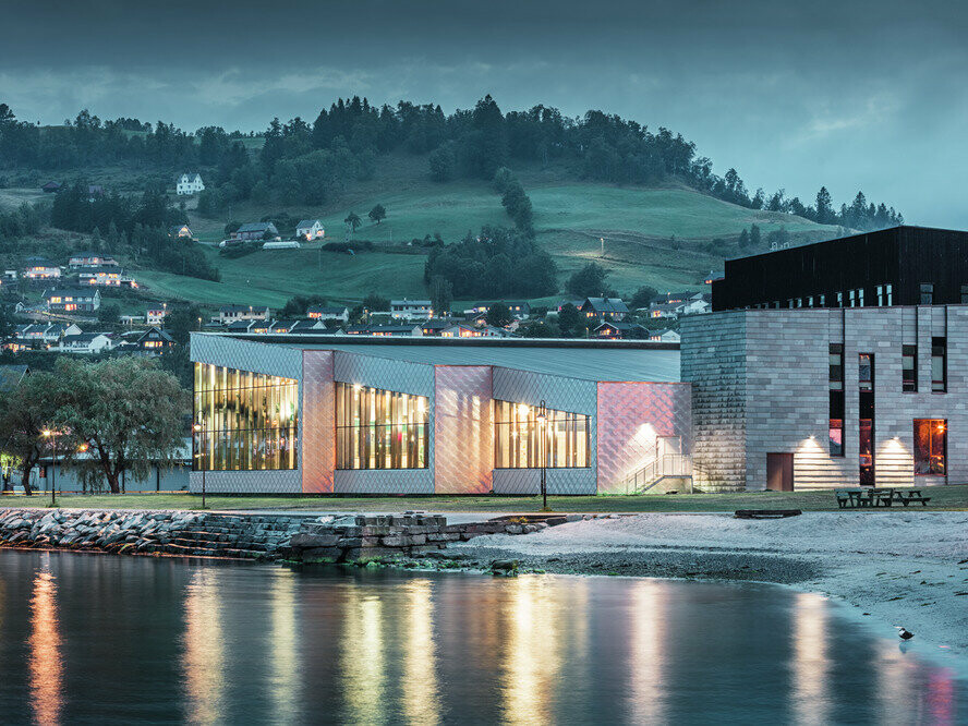 Auf dem Bild ist das Mehrzweckgebäude vom Hardangerbadet Healthcare Center in Oystese vom Wasser aus zu sehen. Diese wurde mit der Wandraute 44x44 in Naturblank eingedeckt. Man sieht einen Teil vom Hardangerfjord, das Objekt beleuchtet in Abenddämmerung und im Hintergrund Oystese.