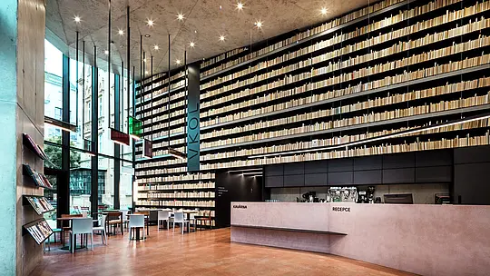 Das Bild zeigt das moderne und stilvolle Innere der Bibliothek in Olmütz, das durch seine beeindruckende Bücherwand dominiert wird. Die bis zur Decke reichenden Regale erzeugen eine dramatische Wirkung, während der schlichte, aber elegante Empfangstresen aus feinstem Beton und Holz eine angenehme Atmosphäre schafft. Besonders hervorzuheben sind die klaren Linien und die präzise Beleuchtung, die das Gesamtbild harmonisch ergänzen. Die Verwendung von PREFABOND Aluminium Verbundplatten und anderen hochwertigen Materialien an der Außenfassade sorgt für eine langlebige und pflegeleichte Gestaltung. Diese Architektur verbindet Funktionalität und ästhetischen Anspruch auf eindrucksvolle Weise.