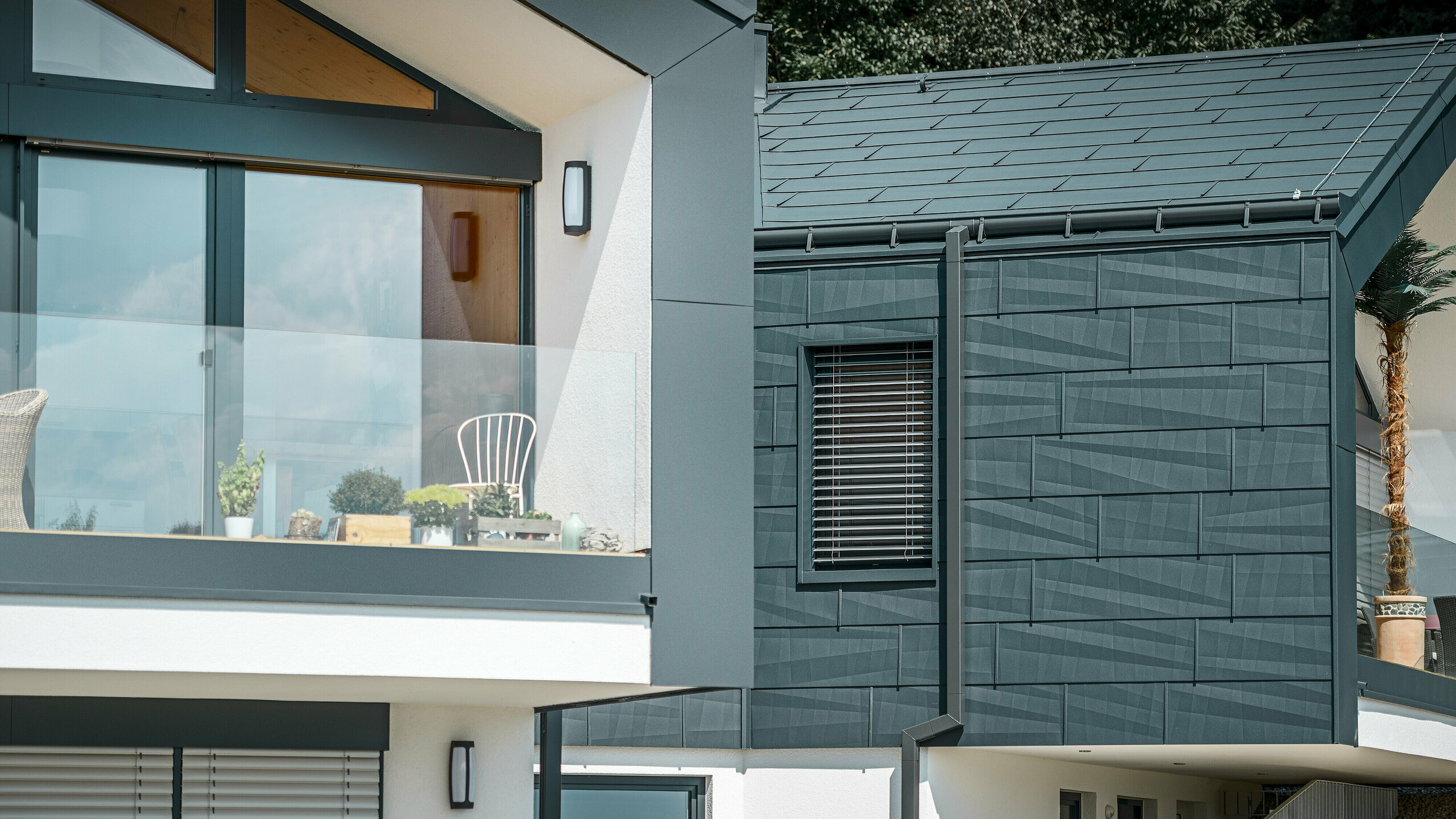 De foto toont een moderne twee-onder-een-kapwoning met een stijlvolle gevel van aluminium. De gevel is uitgevoerd in een donkere antracietkleur en heeft een gestructureerd oppervlak dankzij de PREFA dak- en gevelpaneel FX.12. Op het dak zijn een ardense goot en een vierkante buis van PREFA gemonteerd. Op de bovenste verdieping is een balkon met glazen balustrade te zien, terwijl op de onderste verdieping ramen met lamellenjaloezieën zijn aangebracht. De architectuur oogt strak en minimalistisch, en op de achtergrond zijn bomen te zien die een natuurlijke sfeer creëren.