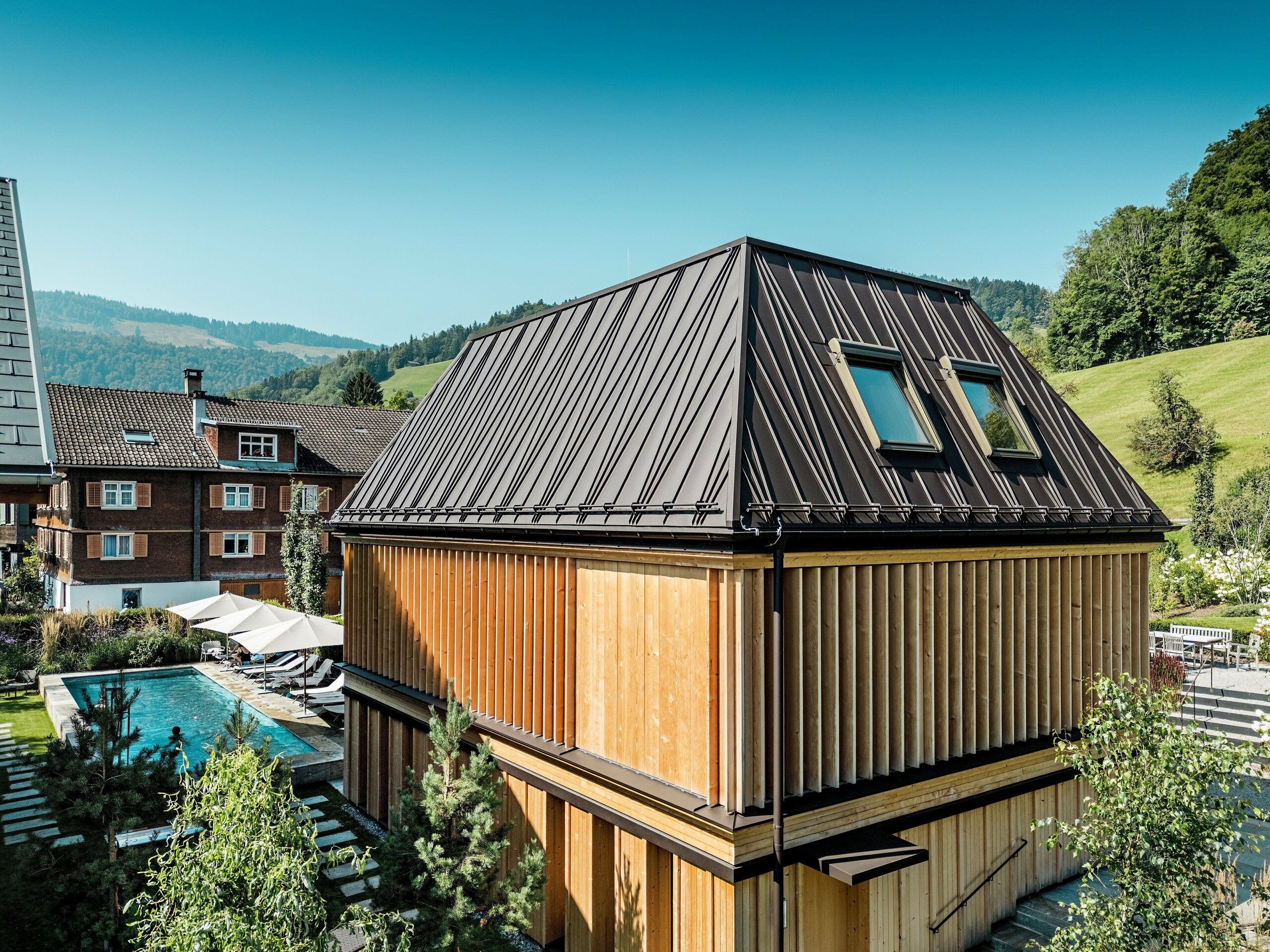 Das moderne „Badehaus“ des Hotel Hirschen in Schwarzenberg an einem sonnigen Tag, eingebettet in die grüne Hügellandschaft des Bregenzerwaldes. Das Gebäude beeindruckt durch sein auffälliges PREFA Stehfalzdach in P.10 Nussbraun mit markantem, schräg verlaufendem Falzmuster und zwei Dachfenstern. Die Fassade besteht aus geschoßhohen, vertikalen Holzlamellen, die Licht, Schatten und Sichtschutz flexibel steuern. Im linken Bildbereich ist der Außenpool mit Liegebereich sichtbar – ein Ort der Ruhe und Erholung. Die Verbindung von hochwertiger Architektur, regionalen Materialien und durchdachter Gestaltung fügt sich harmonisch in die traditionelle Umgebung ein.