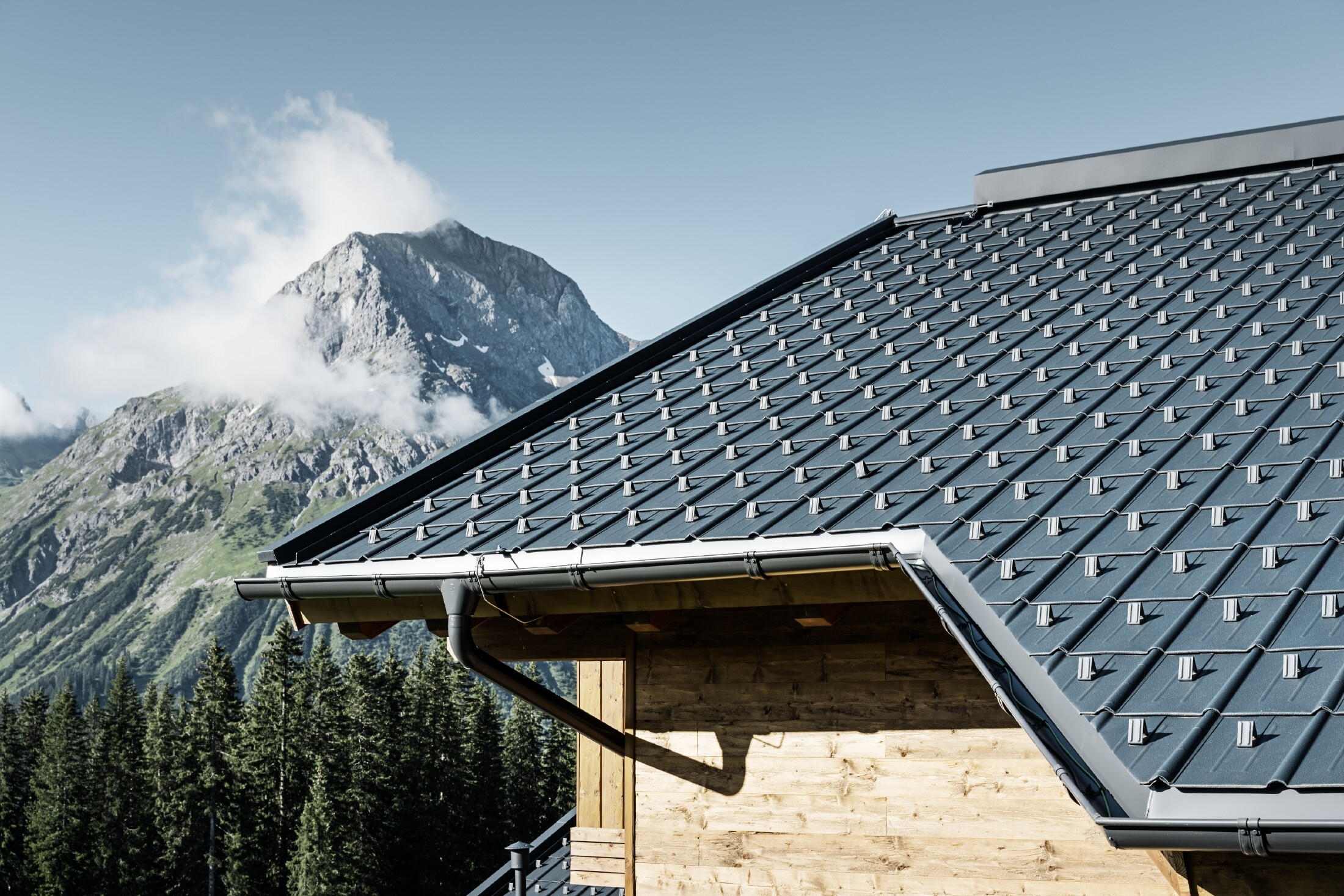 PREFA-dakpan in een bergstreek - dakplaat in antraciet, gemonteerd op de berghut in Lech am Arlberg met bergen op de achtergrond; PREFA-dakgoot in antraciet; houten gevel