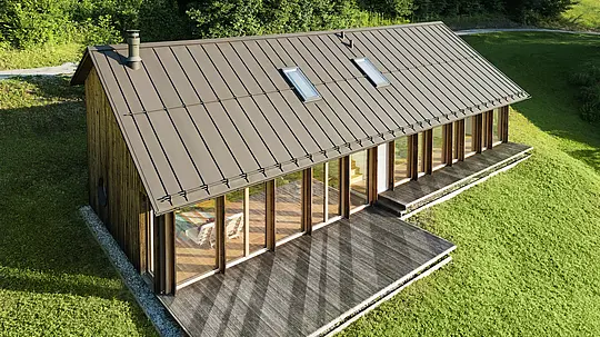 Ein modernes Holzhaus mit großzügiger Glasfassade und einem robusten PREFALZ-Aluminiumdach in der Farbe P.10 Braun – ideal für Hanglagen wie in Vinje. Die hochwertige Dachdeckung garantiert maximale Witterungsbeständigkeit, lange Lebensdauer und eine elegante Optik. Präzise gesetzte Stehfalze und integrierte Dachfenster schaffen ein luftiges und nachhaltiges Wohnkonzept. Die präzise Verarbeitung der Aluminium-Stehfalze wird aus der Vogelperspektive sichtbar.