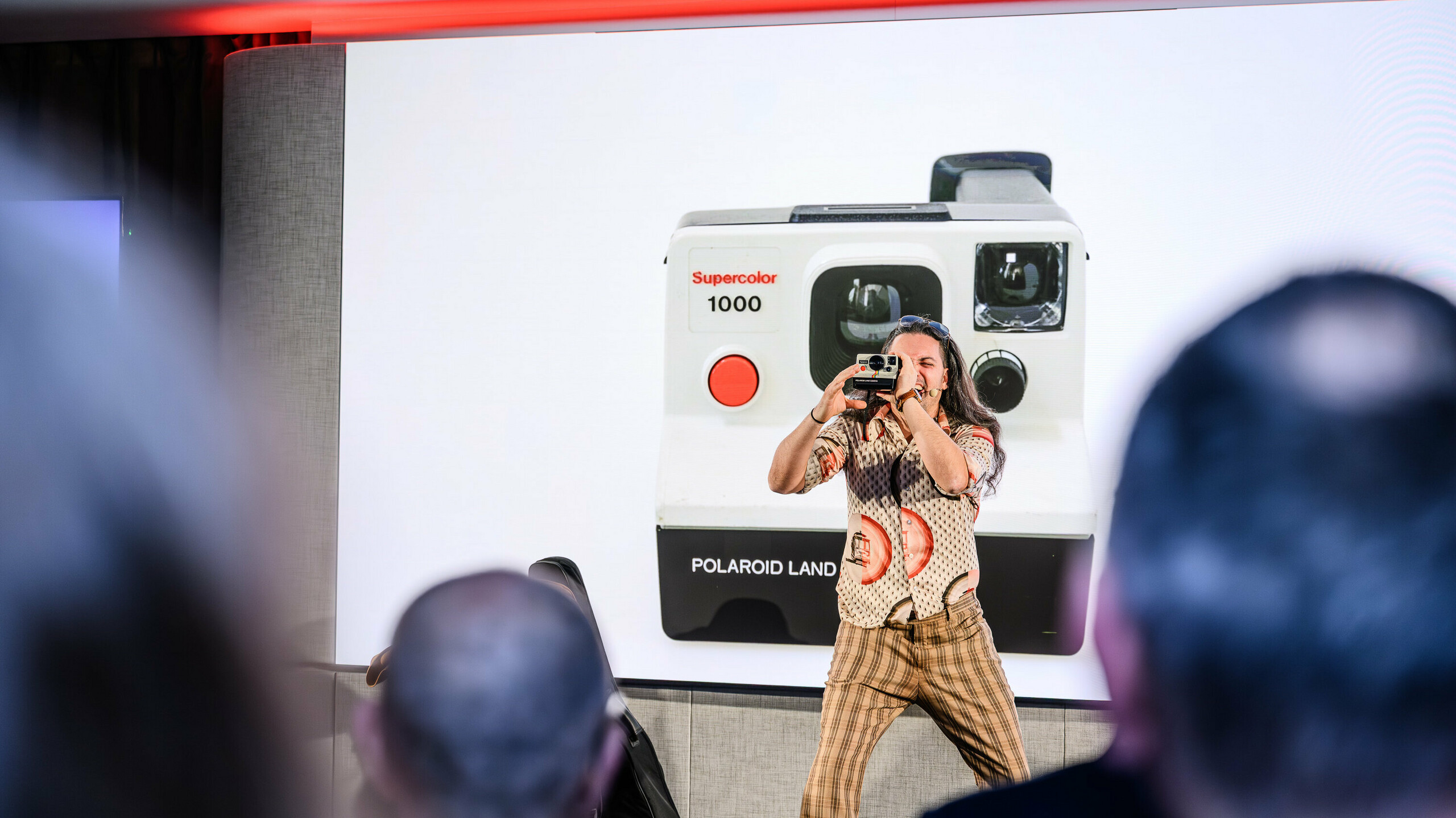 Kreativer Moment bei der PREFARENZEN Präsentation 2026: Der unterhaltsame Moderator fängt mit einer Retro-Polaroidkamera das Publikum ein – vor einer überdimensionalen Projektion der ikonischen Supercolor 1000. Der künstlerische Beitrag schlägt die Brücke zwischen Vergangenheit und Zukunft, genau wie PREFA mit seinen langlebigen Aluminiumprodukten. Ein Abend voller Emotion, Innovation und ästhetischer Impulse.