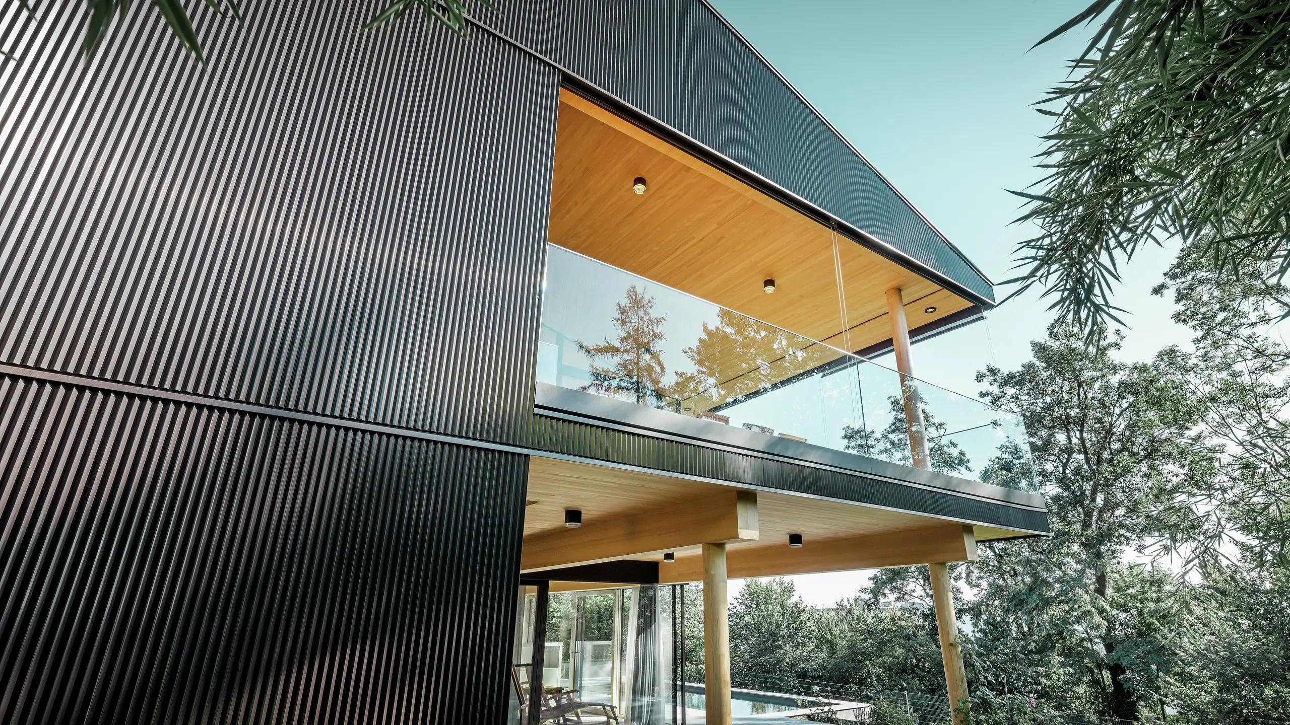 Zijaanzicht van een vrijstaand huis, de gevel is bekleed met donker PREFA-kartelprofiel. Te zien zijn het terras en een ruim balkon. Het plafond of de onderkant is uitgevoerd met houten panelen.