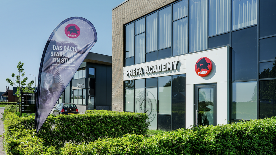 Außenansicht der PREFA Schulungszentrum in Nijkerk mit Werbefahne „Stark wie ein Stier“ und PREFA Logo über der Eingangstür.
