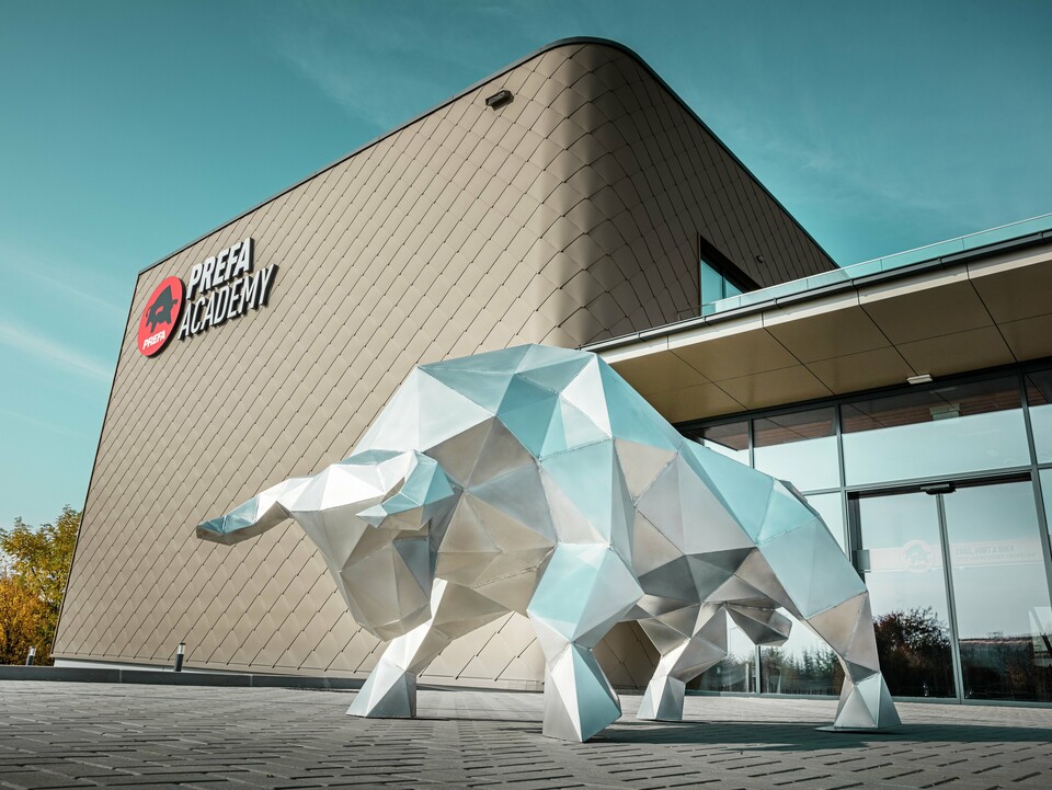 Eine imposante Stierskulptur aus 2 mm blankem Aluminium prägt den markanten Eingangsbereich des neuen PREFA Standortes in Törökbálint. Die moderne Fassade mit PREFA Wandrauten 44 × 44 in Bronze verleiht dem neu errichteten Bauwerk eine elegante, witterungsbeständige und langlebige Außenhülle. Die Glasschiebetüren sorgen für eine lichtdurchflutete Atmosphäre beim Betreten des Gebäudes. Das gut sichtbare PREFA Academy Logo an der Fassade unterstreicht den starken Charakter des Firmengebäudes.