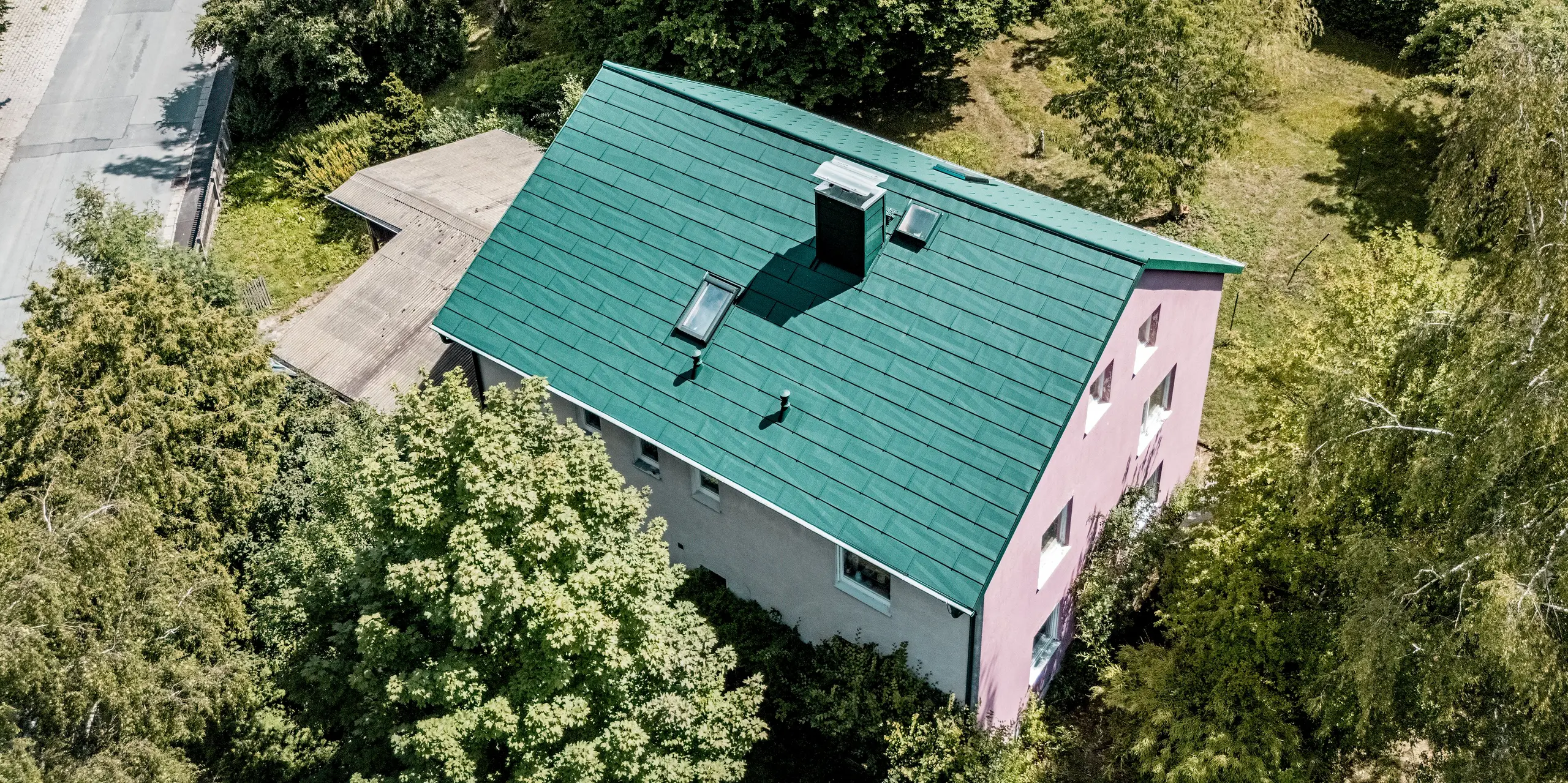Luftaufnahme eines sanierten Einfamilienhauses in Schwarzenbach-Martinlamitz mit PREFA Dachpaneel FX.12 in P.10 Moosgrün. Das moderne Aluminiumdach beeindruckt durch seine klare Struktur, das integrierte Dachfenster und den stilvoll verkleideten Kamin. Besonders auffällig ist der farbliche Kontrast zwischen dem moosgrünen Dach und der violetten Giebelfassade. Umgeben von sattem Grün fügt sich das Haus harmonisch in die Landschaft ein. Das PREFA Dach überzeugt durch Langlebigkeit, Sturmfestigkeit und eine wartungsfreie Oberfläche – ideal für nachhaltige Sanierungen.
