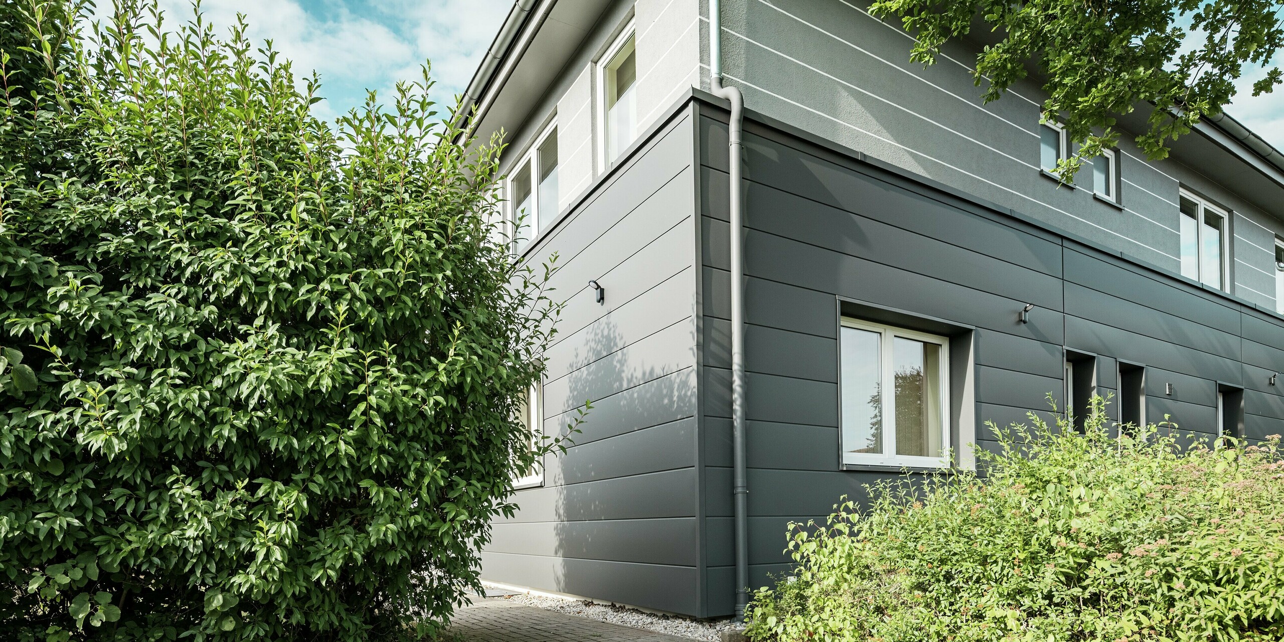 Die PREFA Sidings in der Farbe P.10 Dunkelgrau prägen die Fassade der Kaminkehrer-Innung in Bindlach und verleihen ihr eine moderne, klar strukturierte Optik. Die Aluminium-Paneele sehen nicht nur gut aus, sondern bieten auch langlebigen Schutz vor Witterungseinflüssen. Das Bild zeigt die harmonische Verbindung zwischen der schlichten Fassadengestaltung und der grünen Bepflanzung im Vordergrund. Die pflegeleichten und widerstandsfähigen Paneele sind eine nachhaltige Lösung für eine moderne Fassadengestaltung.