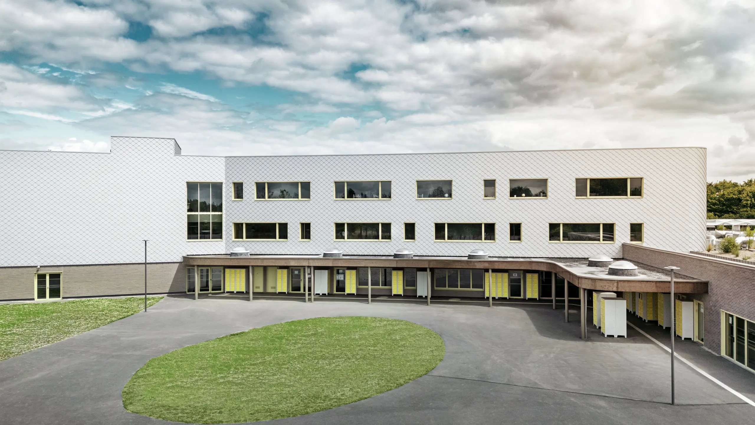 Die Rückseite des Collège Léontine Dolivet mit seinem weitläufigen Schulhof und der klaren Architektur. Die Fassade besteht aus silbermetallischen PREFA-Wandelementen, die dem Gebäude ein modernes und elegantes Aussehen verleihen. Mehrere Fensterreihen und gelbe Türen gliedern die Struktur, während der gepflasterte Innenhof mit seiner zentralen Grünfläche viel Platz zum Entspannen bietet. Die robuste Aluminiumfassade vereint Leichtigkeit, Witterungsbeständigkeit und ansprechendes Design – ein funktionales und ästhetisch attraktives Gebäude in Cesson-Sévigné bei Rennes.