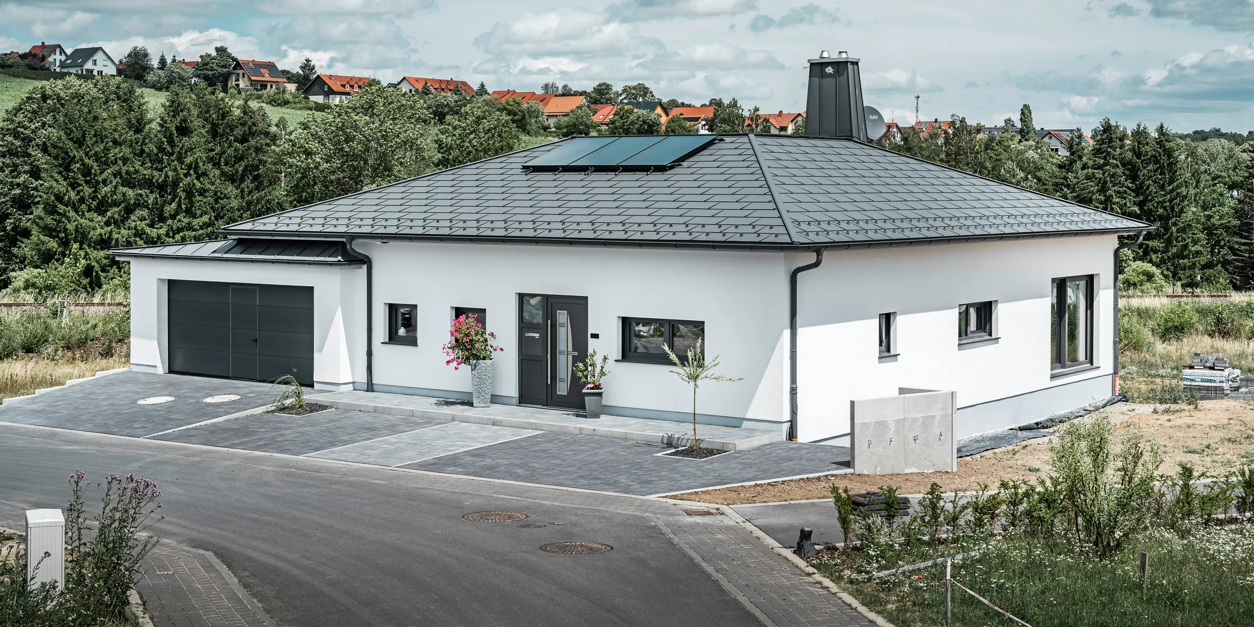 Elegante eengezinswoning in Ilmenau, Thüringen, met een hoogwaardig PREFA-dak in de kleur P.10 donkergrijs, voorzien van de robuuste dakpan R.16. De garage sluit qua kleur en stijl aan door het gebruik van PREFALZ. De blikvanger van het pand is het functioneel ontworpen atrium en de met de PREFA-stier versierde schoorsteen, die het moderne design en de innovatieve daktechnologie van PREFA onderstrepen. De zonnepanelen passen naadloos in het duurzame concept van het huis. Het doordachte totaalconcept van de bungalow wordt gecompleteerd door het PREFA-dakafvoersysteem, dat eveneens in donkergrijs is uitgevoerd.