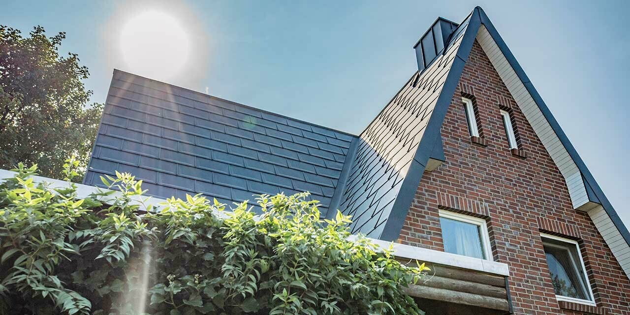 Zadeldak gedekt met de eenvoudige PREFA aluminium dakpan R.16 in antraciet. De gevel is een rustieke baksteengevel, achter het huis gaat net de zon op.