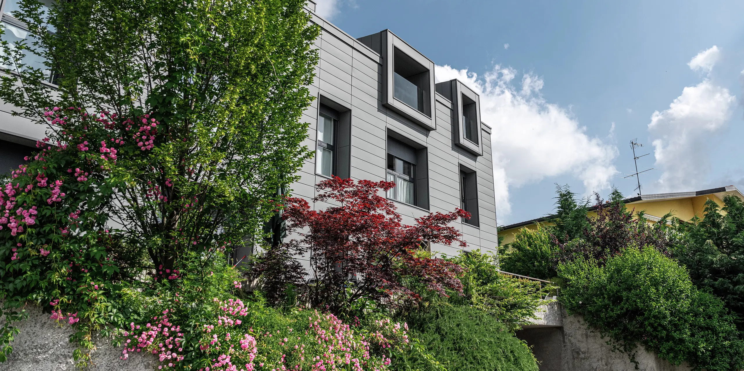Modern wooncomplex in Faloppio, Italië, met stijlvolle gevel van PREFA Sidings. De uitkragende ramen vormen een unieke architectonische blikvanger, terwijl de omringende planten en bloemen de natuurlijke schoonheid van het object onderstrepen. De duurzame aluminium bekleding van PREFA zorgt voor een esthetische en functionele oplossing onder alle weersomstandigheden.